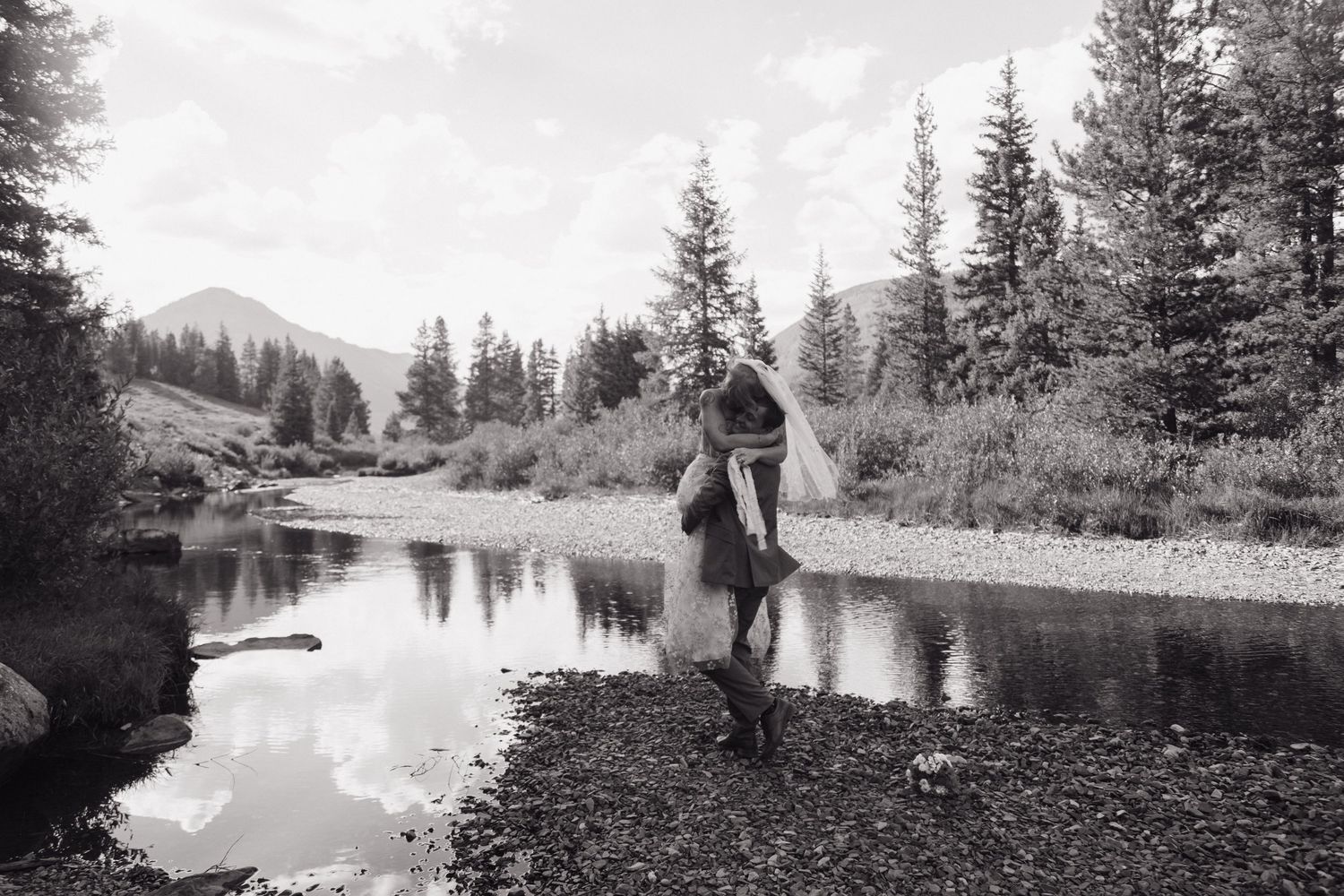 Couple embracing by a reflective mountain stream surrounded by pine trees in a black and white outdoor photo.