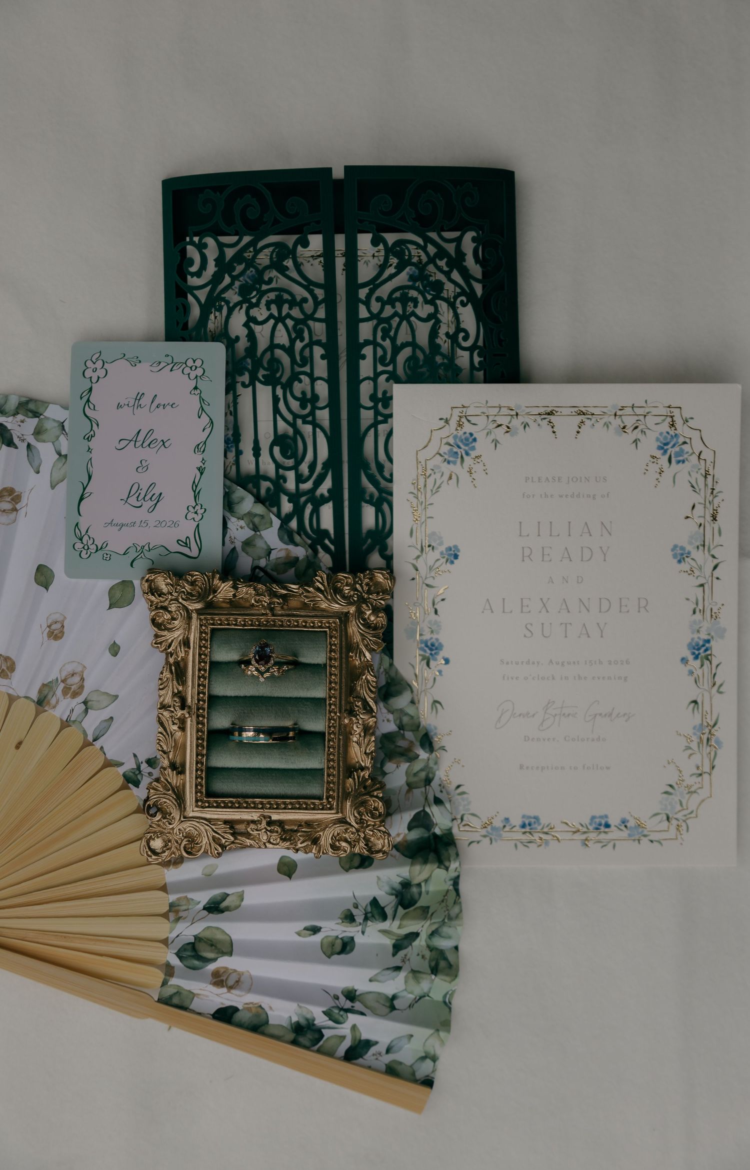 Elegant wedding stationery suite featuring floral invitations, laser-cut green folder, gold frame with rings, and botanical fan.