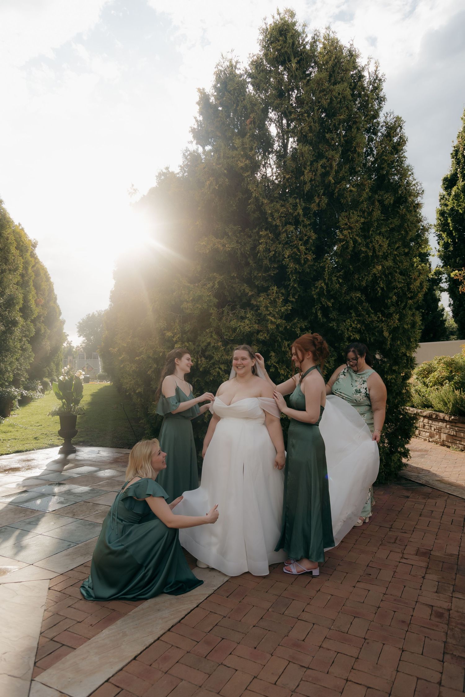 Bridesmaids in sage green dresses help bride with her white gown outdoors near tall evergreen trees in golden sunlight.