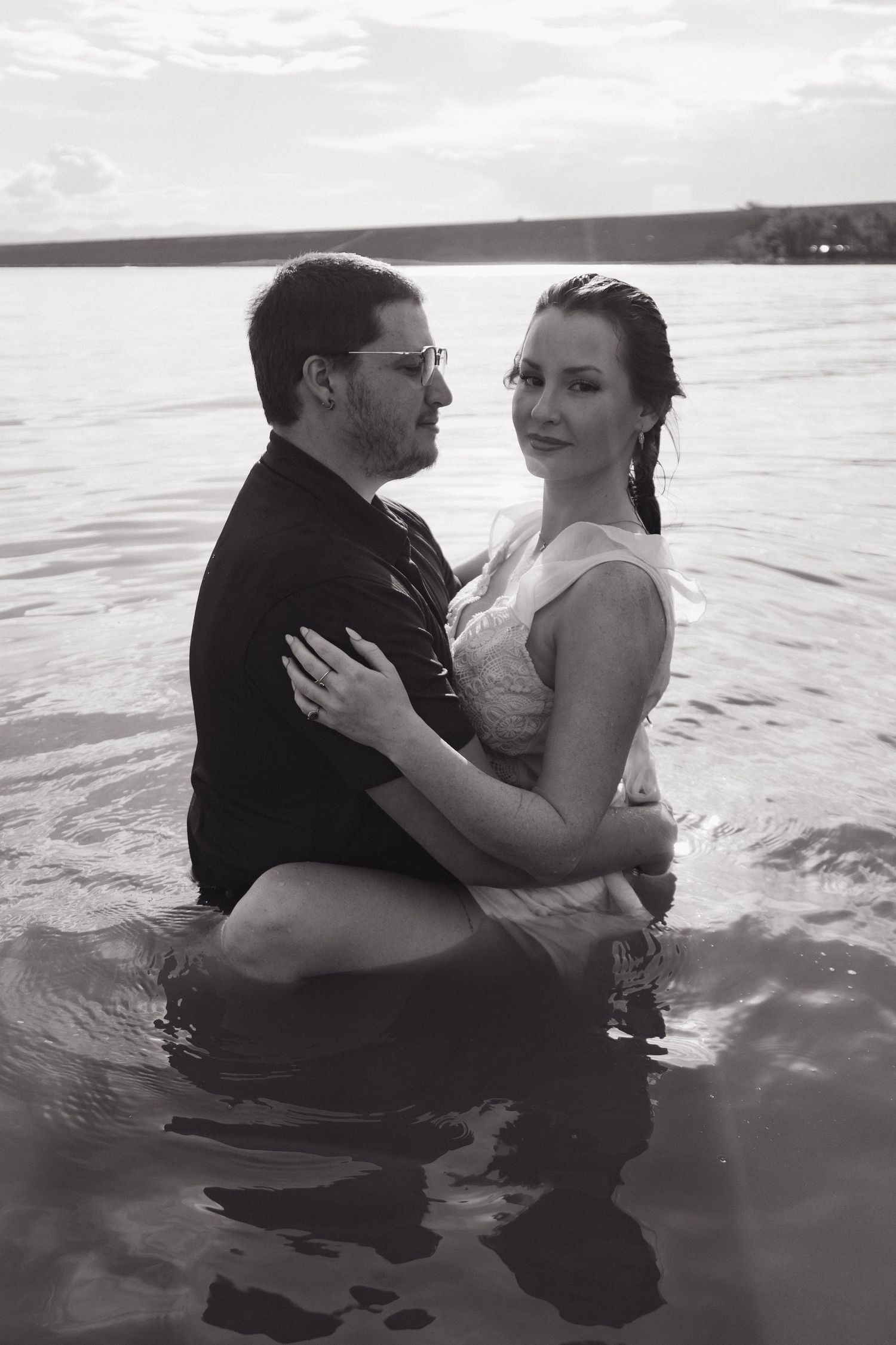 Black and white photo of couple embracing in lake water, woman in wet white dress.