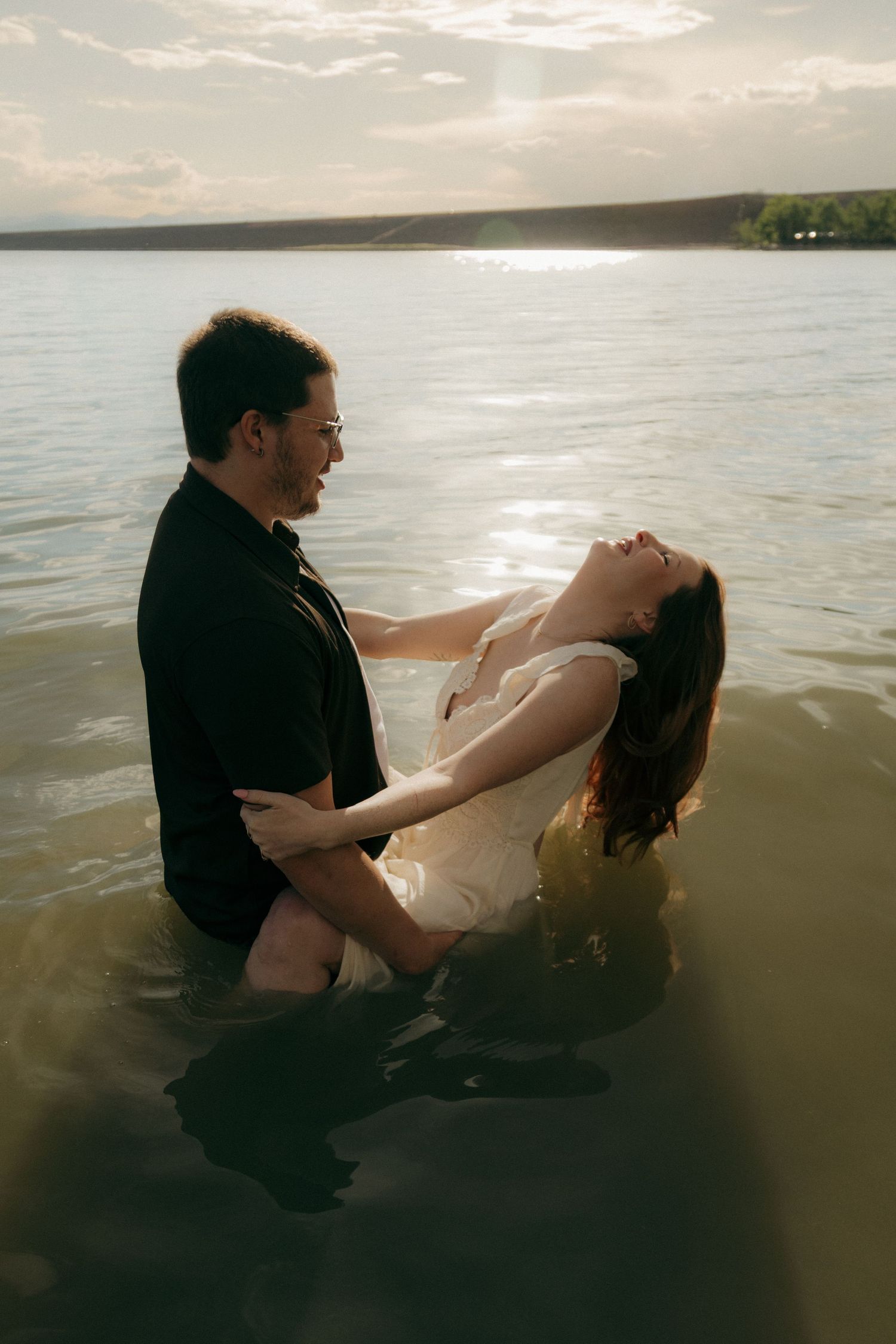 Romantic couple standing in lake water at sunset, woman leaning back in wet white dress.