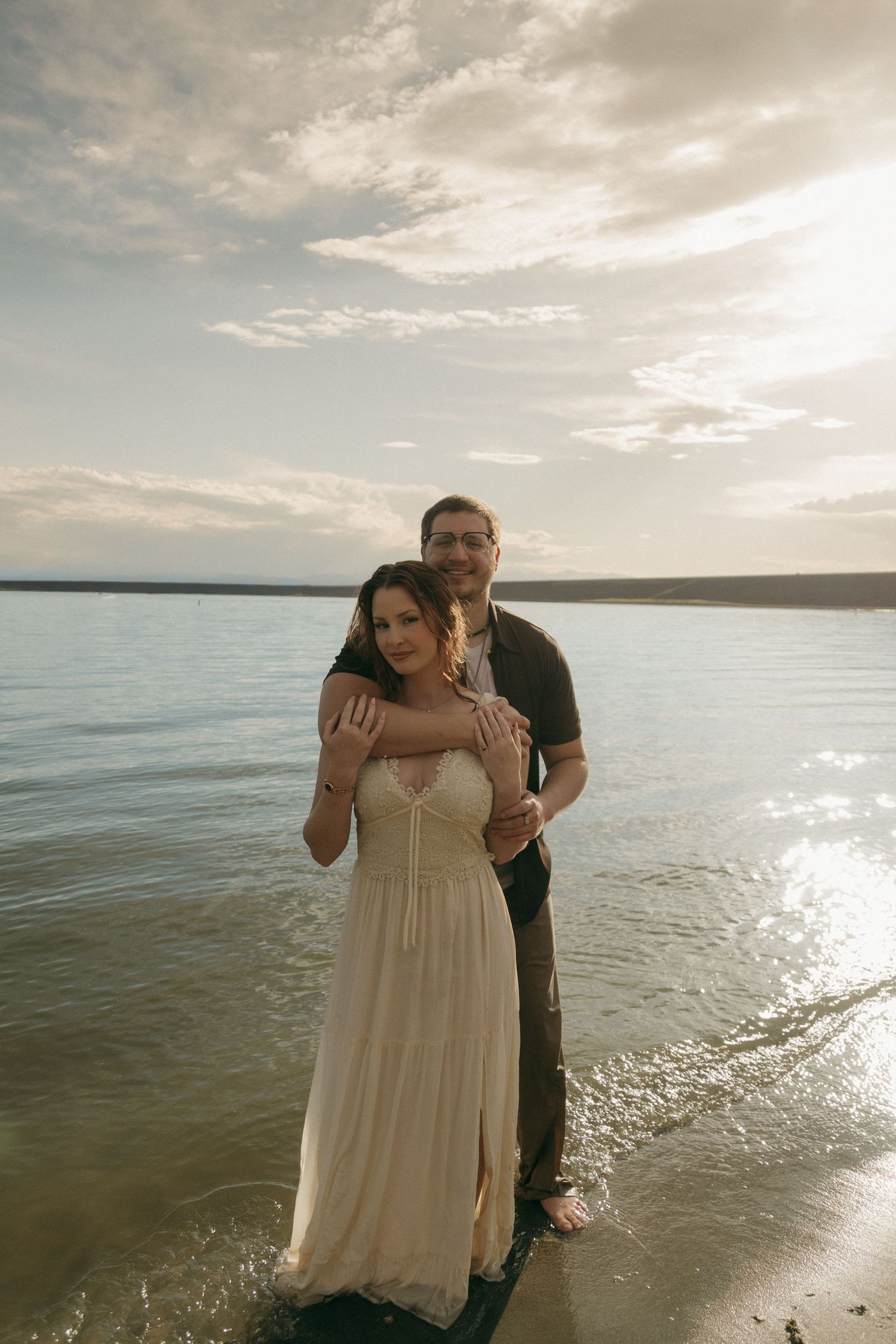 Couple standing in shallow water at sunset, man embracing woman in white dress from behind.