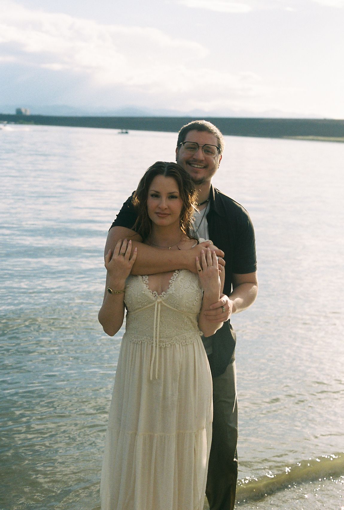 Smiling couple posing together on a lakeside beach, man embracing woman in a white dress.