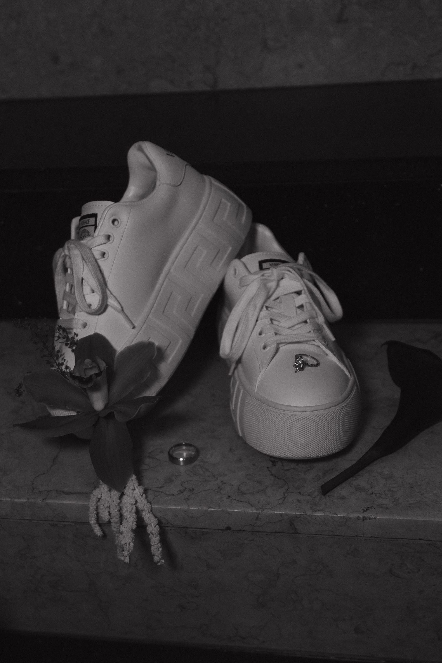 White Versace sneakers with a floral boutonniere and ring on marble surface.