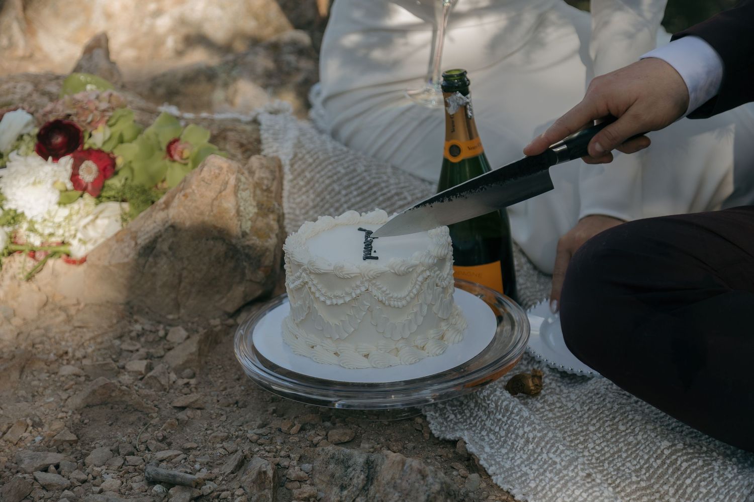 Couple cuts a white wedding cake beside champagne bottle during intimate outdoor celebration.