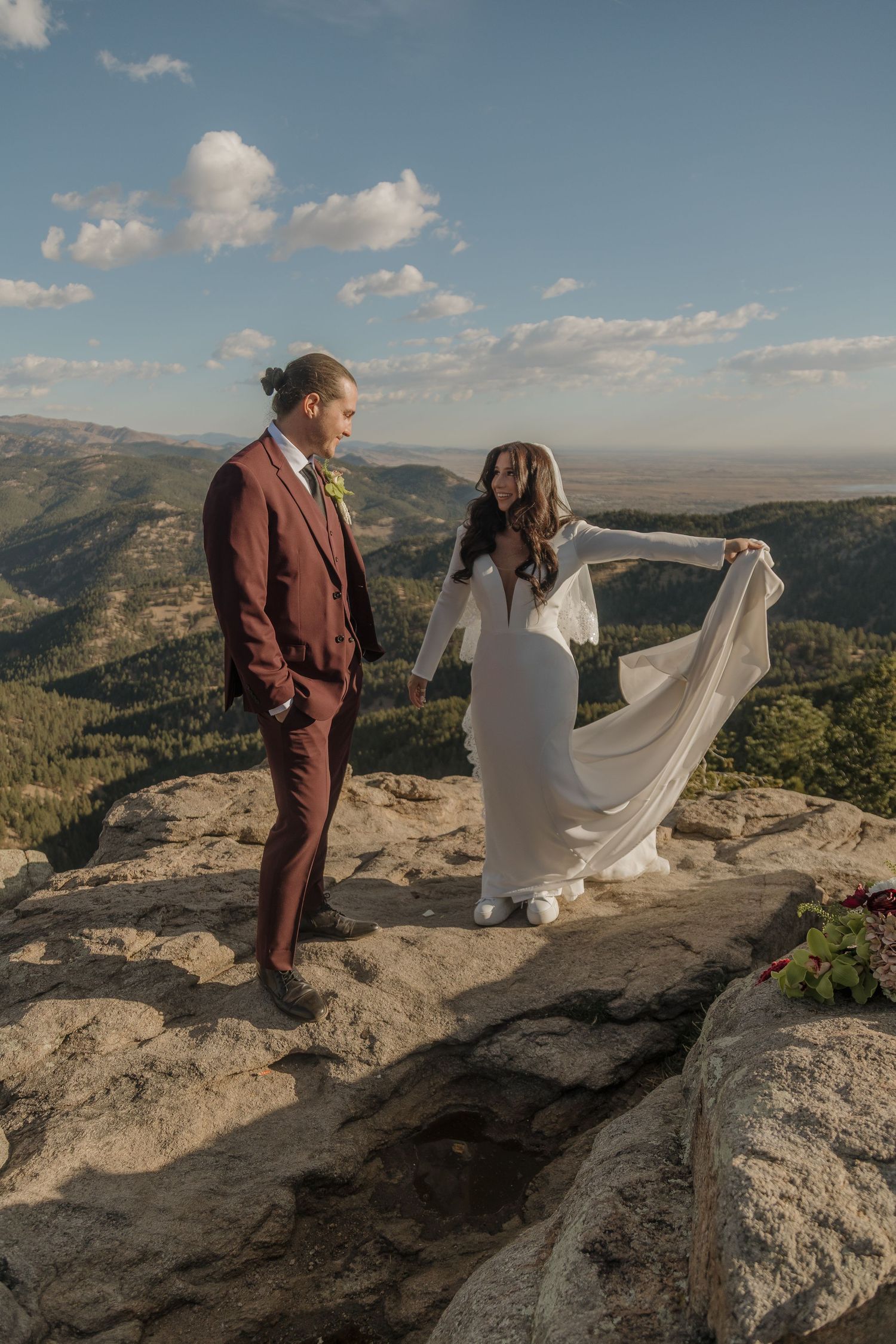Bride in white gown holds flowing veil while groom in burgundy suit stands on a scenic mountain overlook.