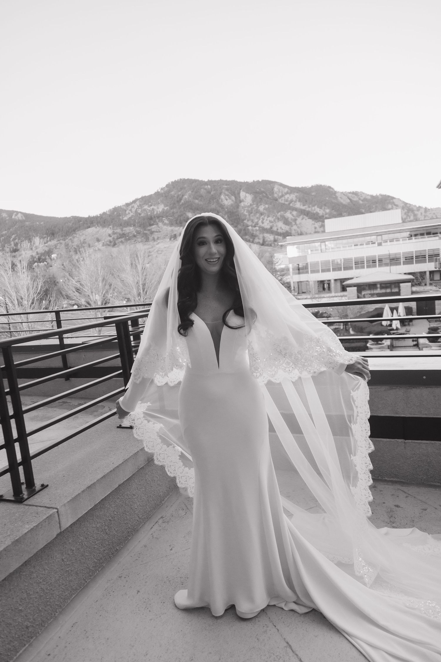 Black and white portrait of bride in fitted gown and lace veil on outdoor terrace with mountains.