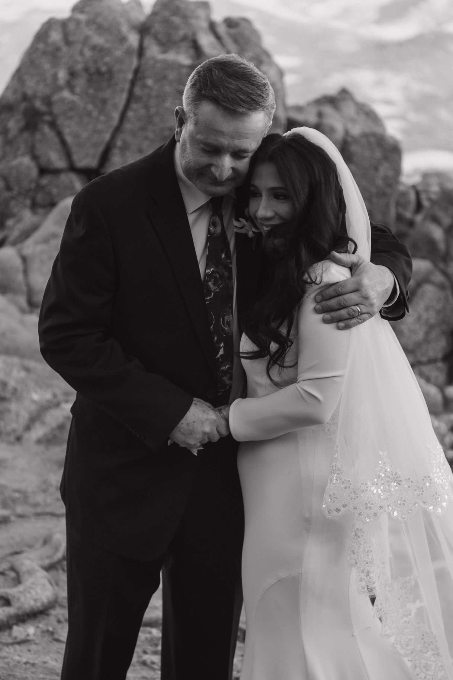Black and white photo of bride and older man sharing emotional embrace on rocky mountain wedding location.