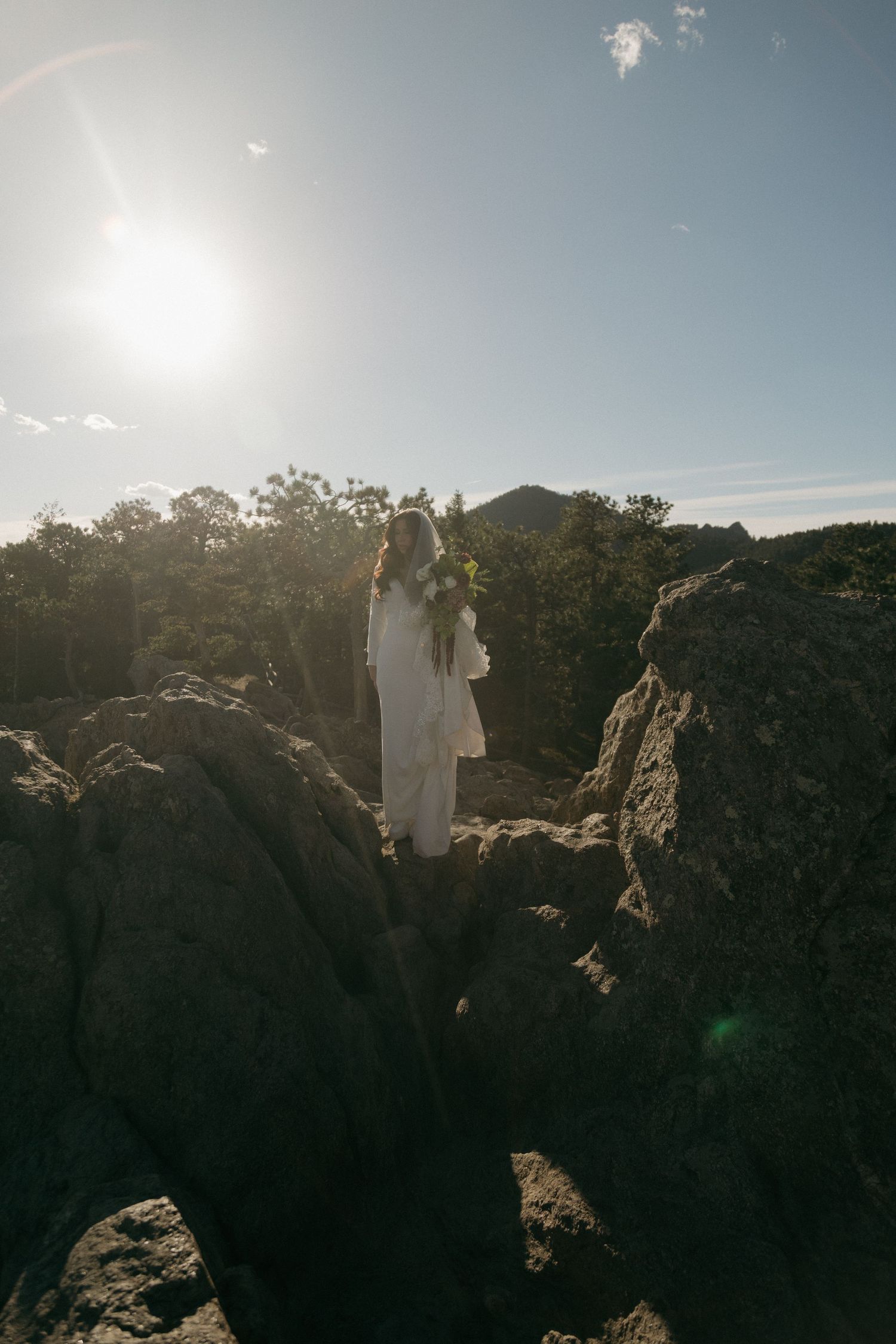 Bride in white dress holding bouquet stands on rocky mountain landscape with sun flare and trees in background.