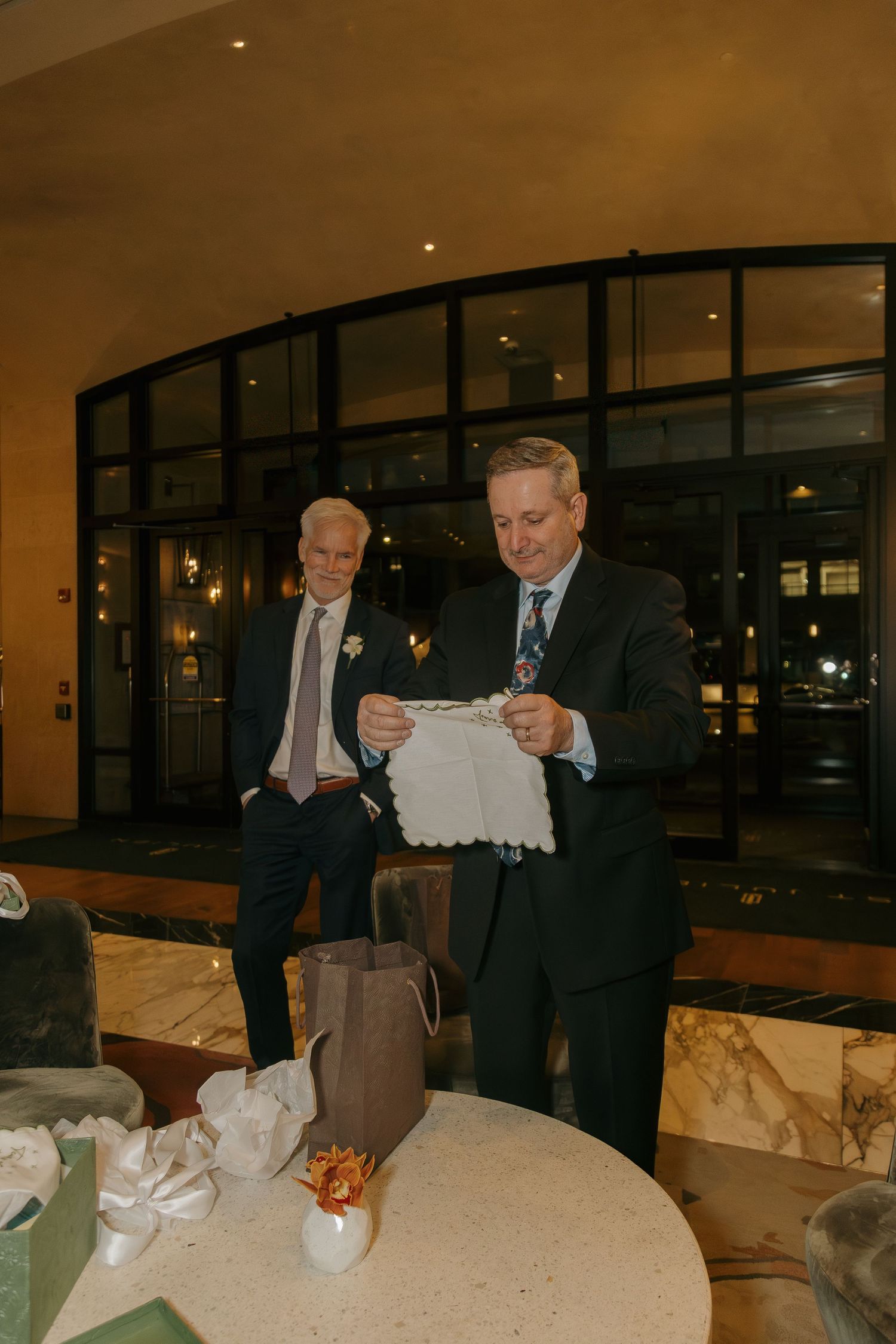 A man in a suit examines a white lace gift at a wedding reception while another guest looks on.