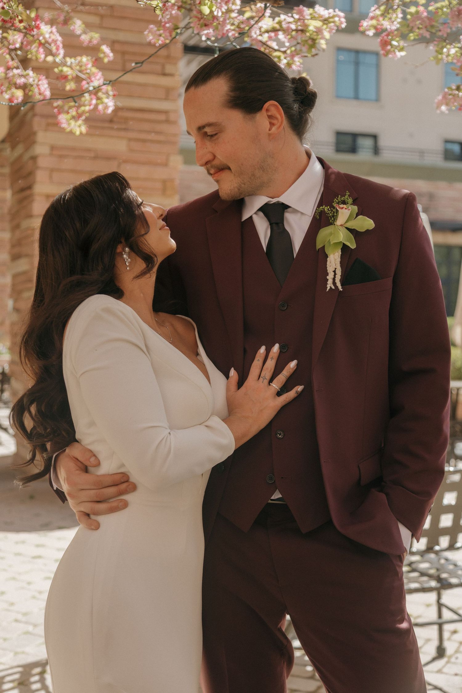 Bride and groom embracing romantically beneath pink cherry blossoms in an urban courtyard.