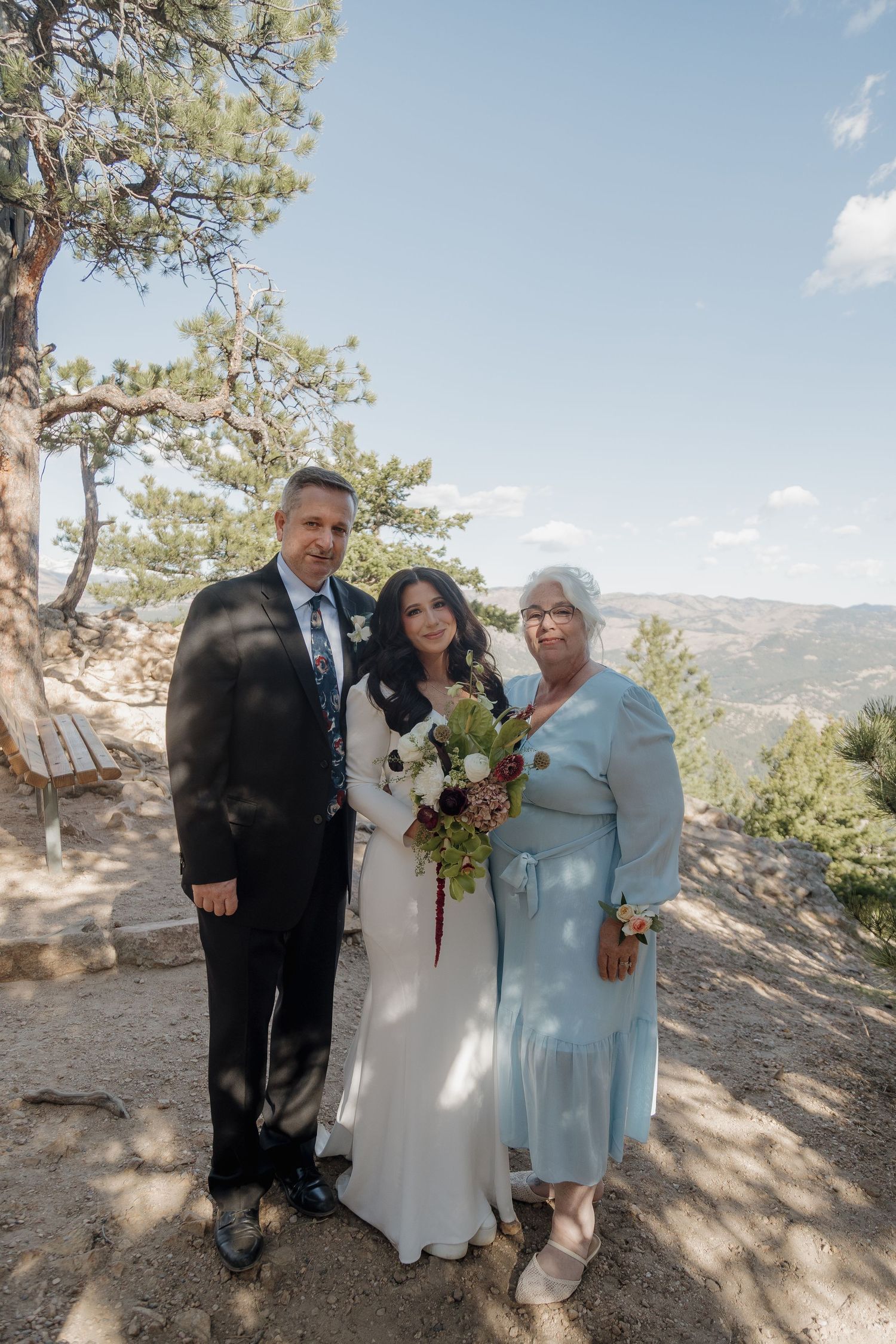 Bride with bouquet poses with two family members on a mountain overlook on wedding day.