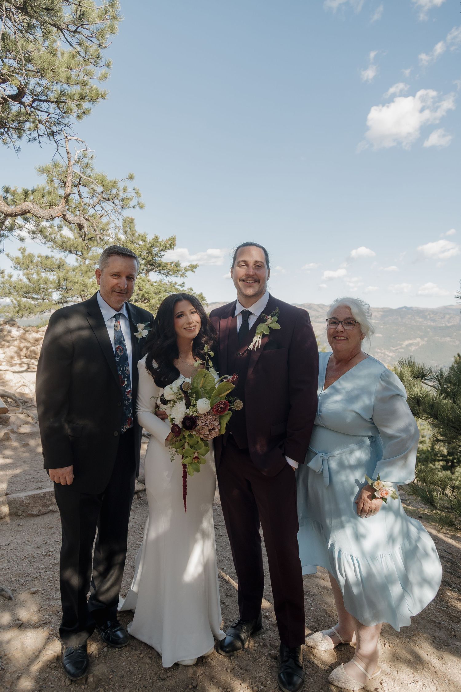 Bride and groom pose with family members holding bouquet under blue sky with mountain views.