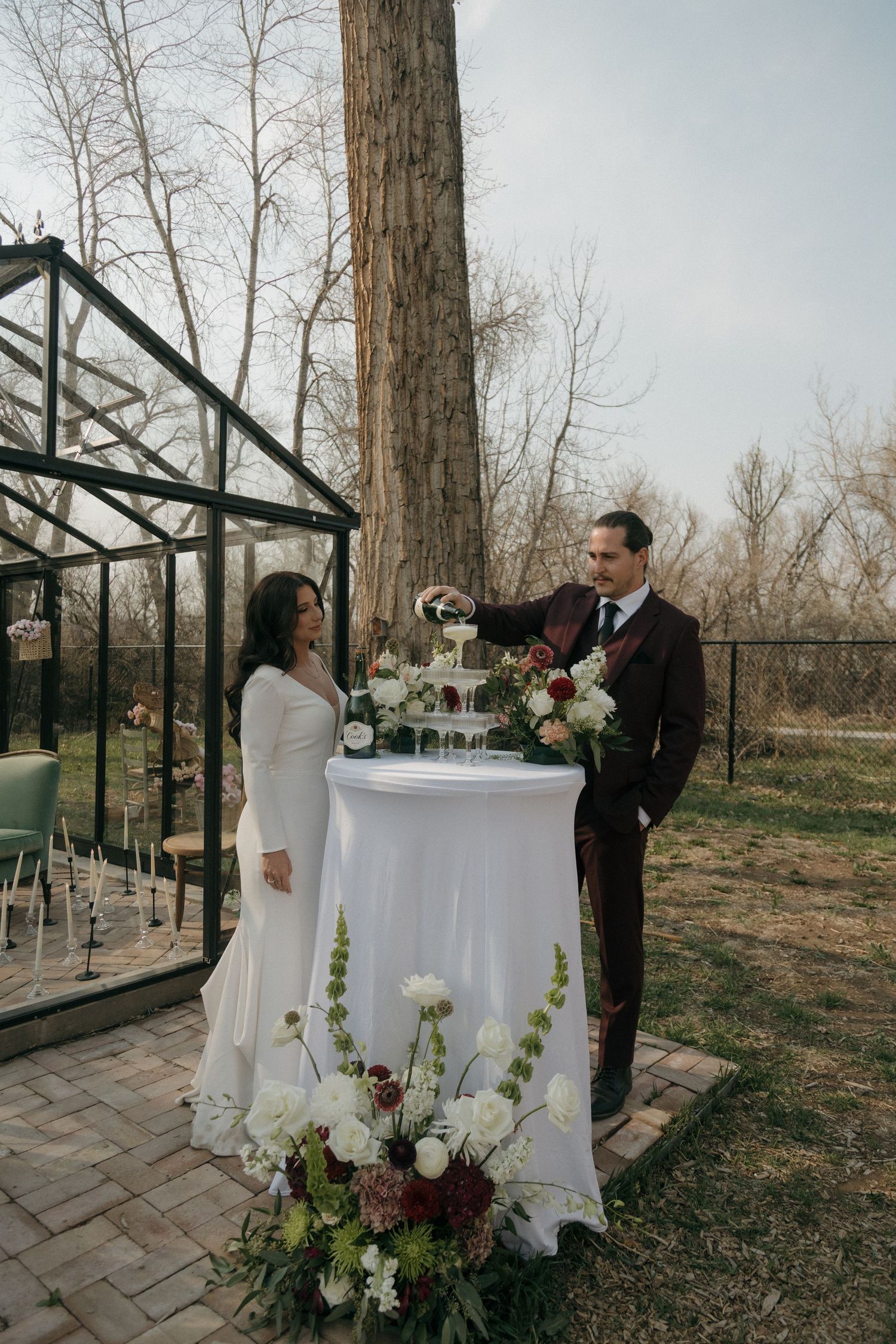 Groom in burgundy suit pouring champagne tower beside bride in white gown at outdoor garden wedding.