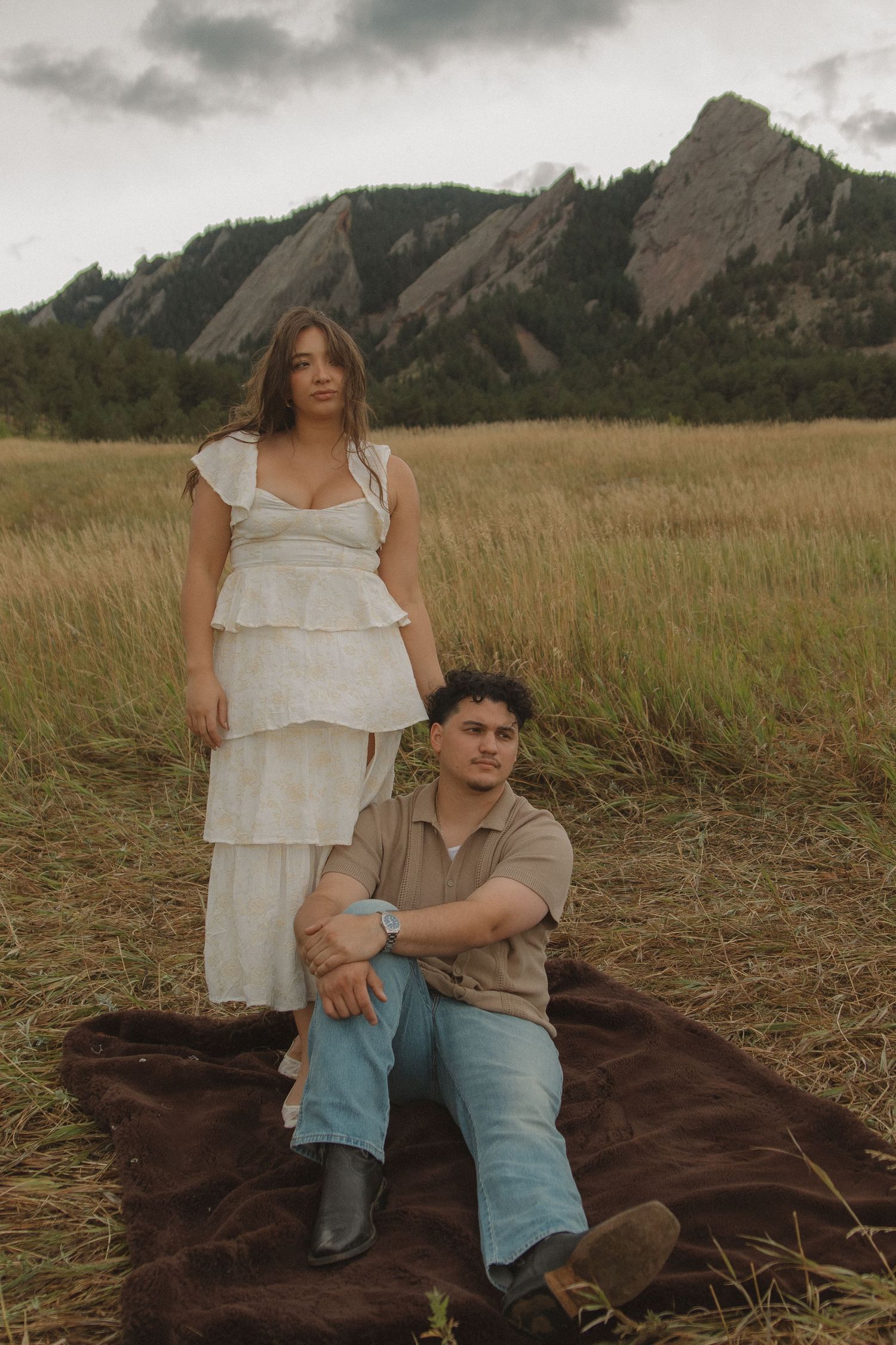 Couple poses in golden field with dramatic Flatirons rock formations behind them near Boulder, Colorado.