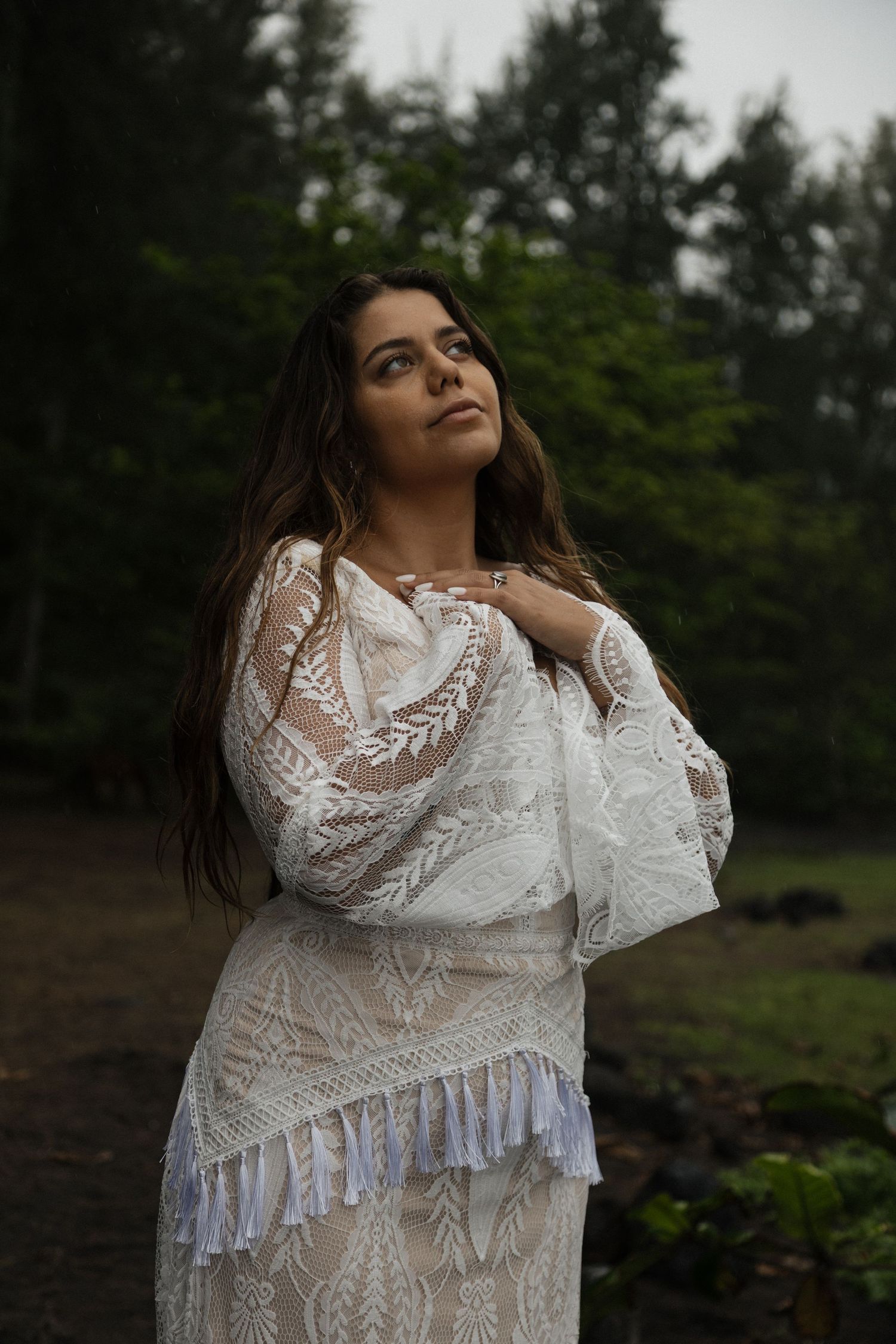 Bride in bohemian white lace wedding dress gazing upward surrounded by tall green forest trees.