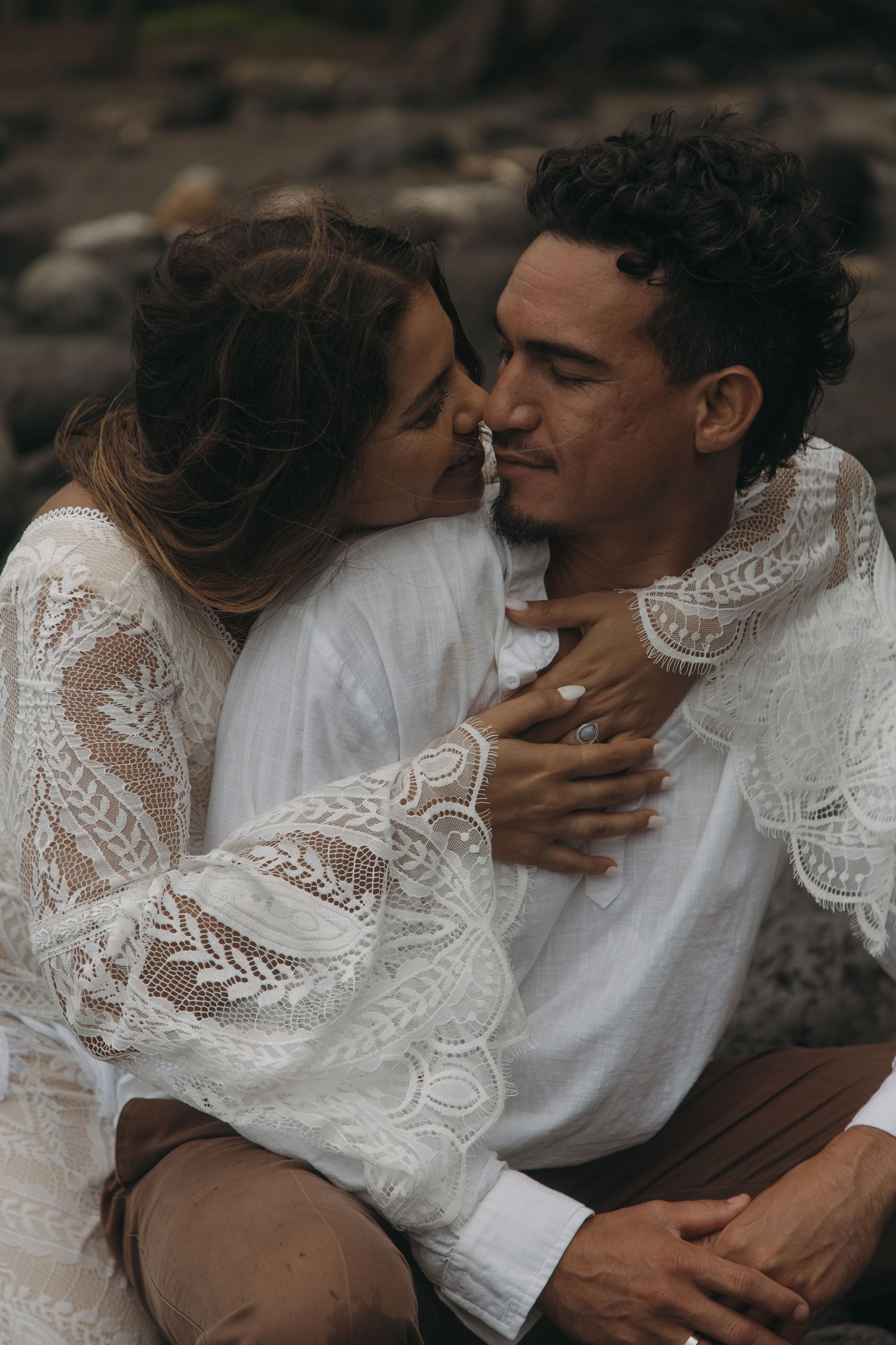 Couple sitting on rocks, bride in lace wedding dress leaning in for a kiss with groom in white shirt.