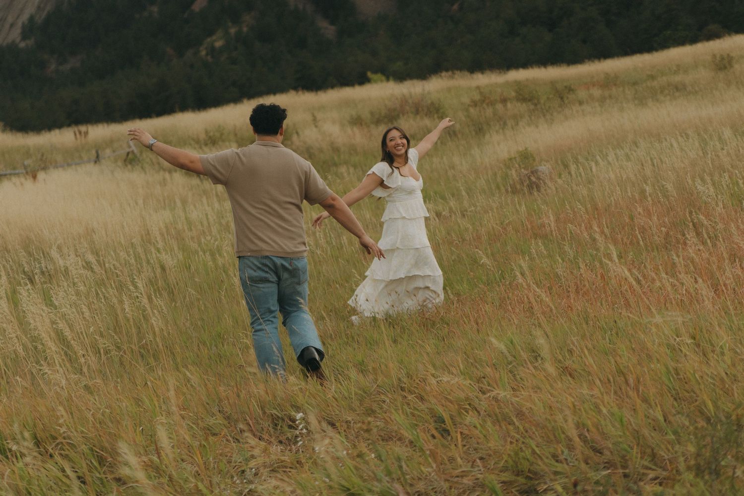 Joyful couple running through golden grass field with mountains in background.