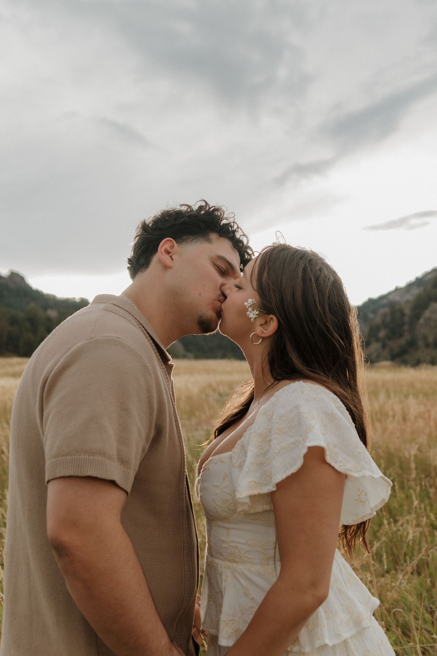 Couple sharing a romantic kiss in a golden field with dramatic cloudy skies and mountain backdrop.