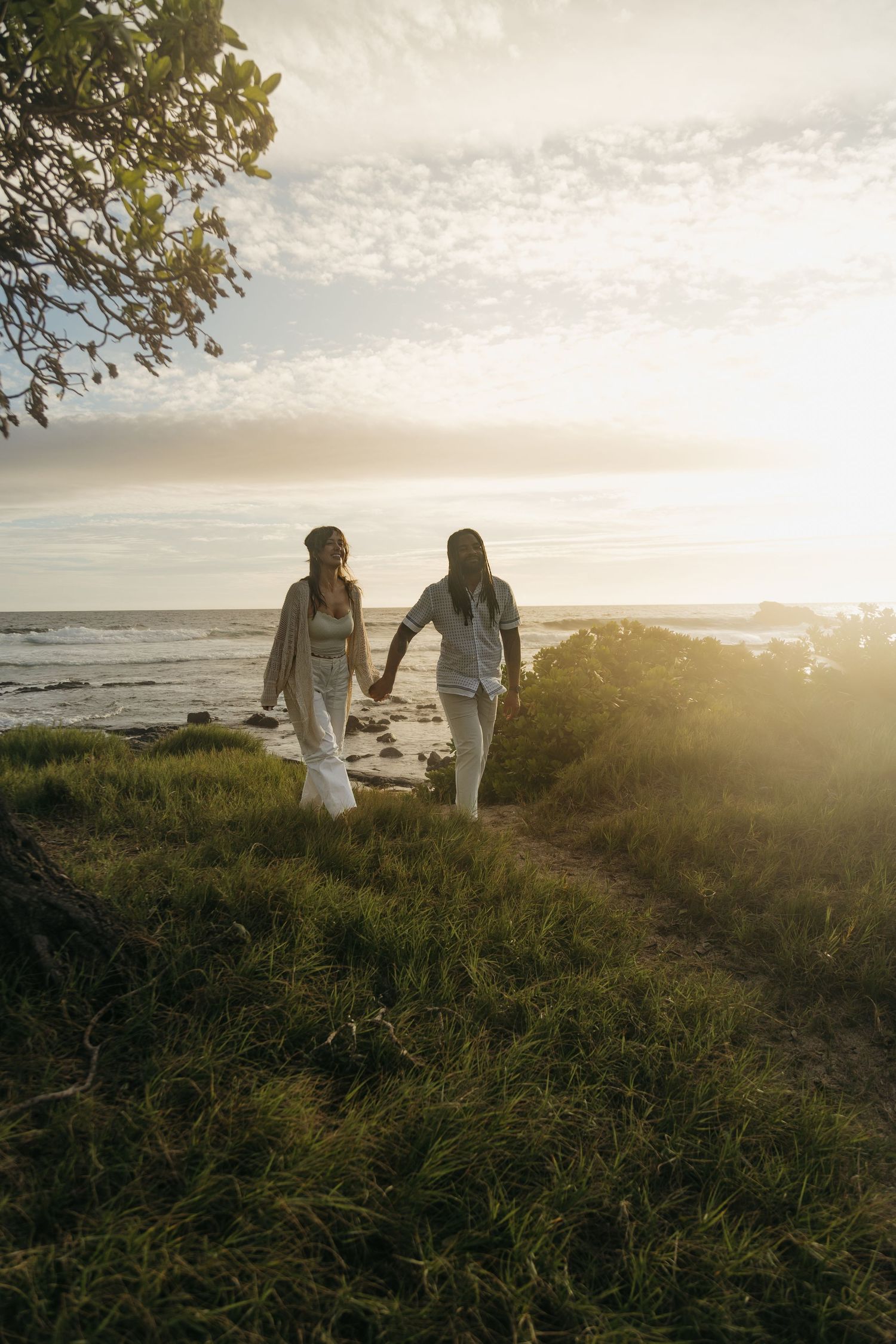 Two women holding hands walk along a grassy coastal path at golden sunset near the ocean.