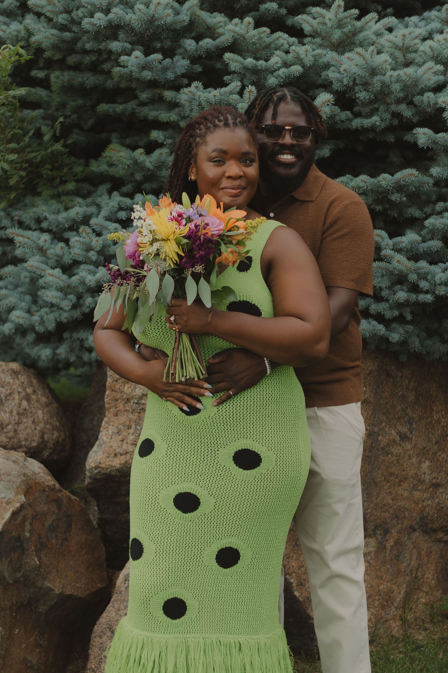 A couple poses outdoors near evergreen trees while wearing a lime green polka dot dress and brown top.