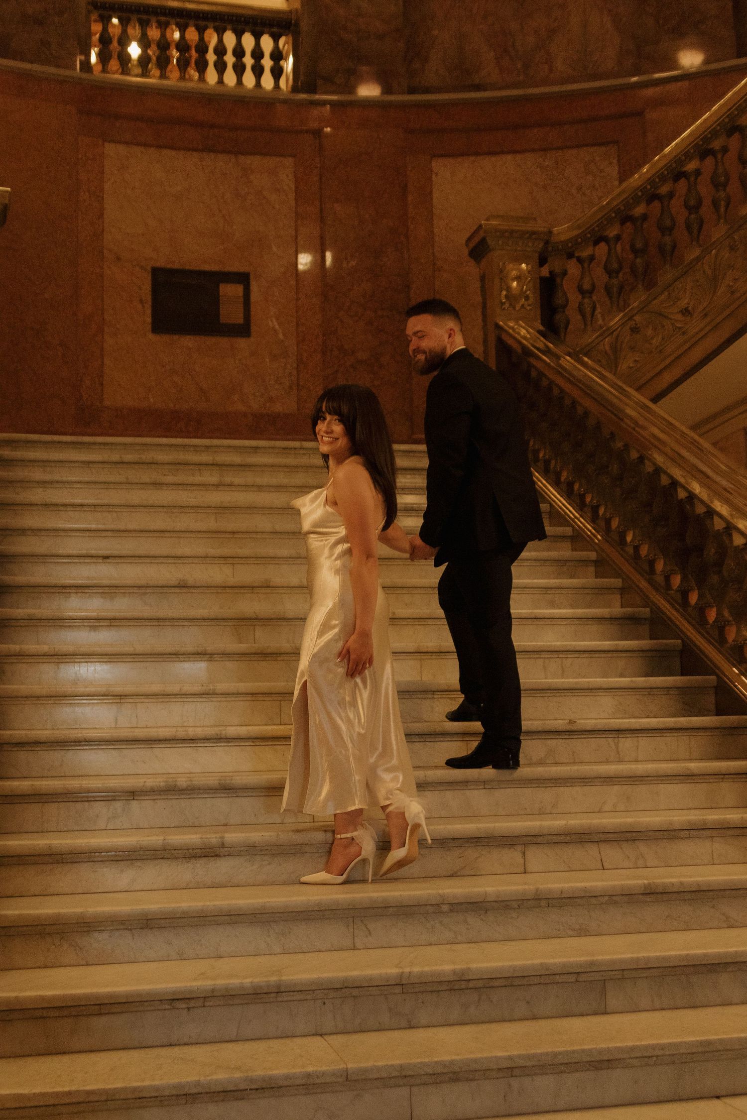 A couple walking down elegant marble stairs in formal attire at a grand venue.