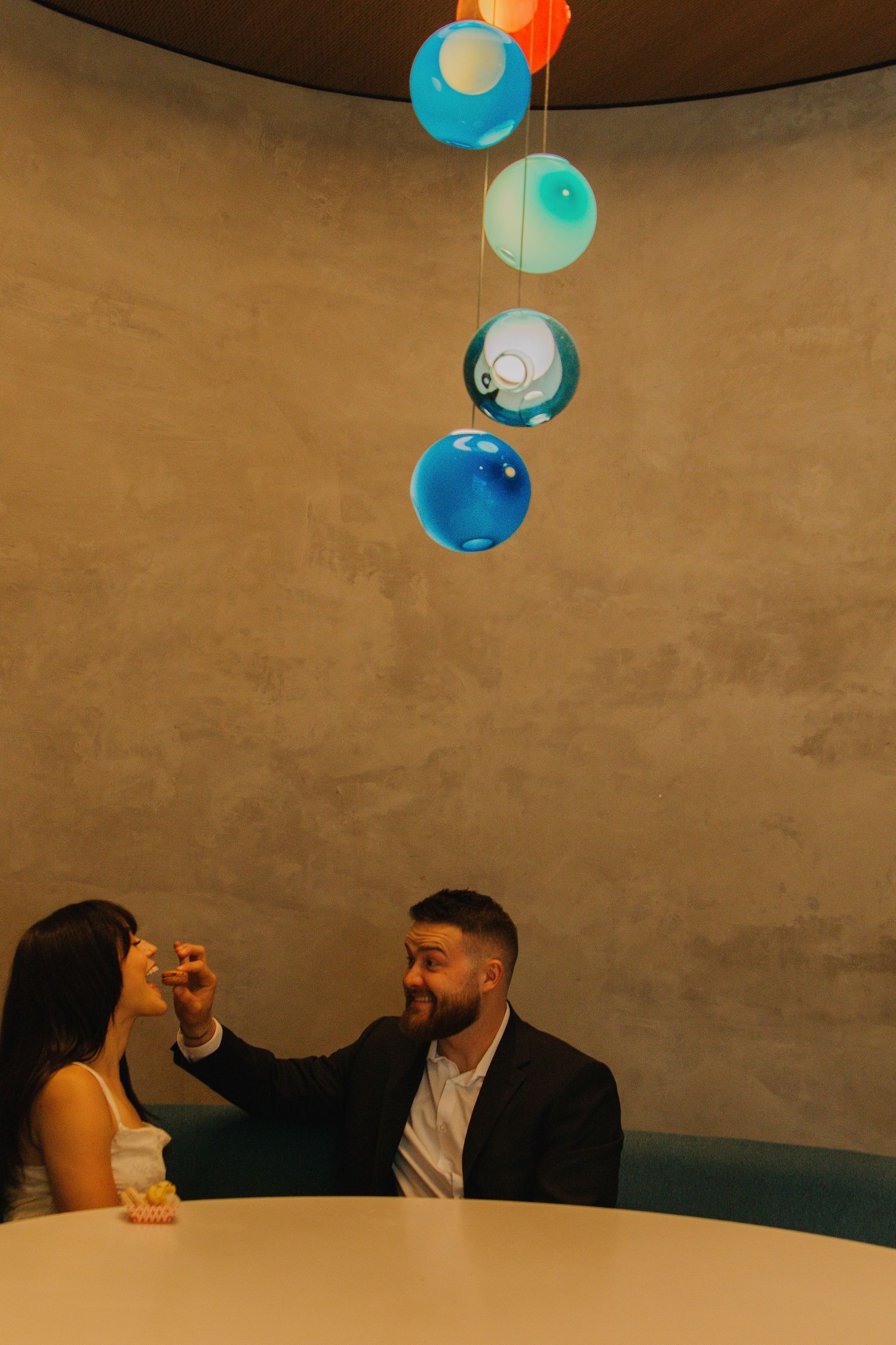 A romantic couple shares an intimate moment at a table under hanging blue spherical lights in a modern restaurant.
