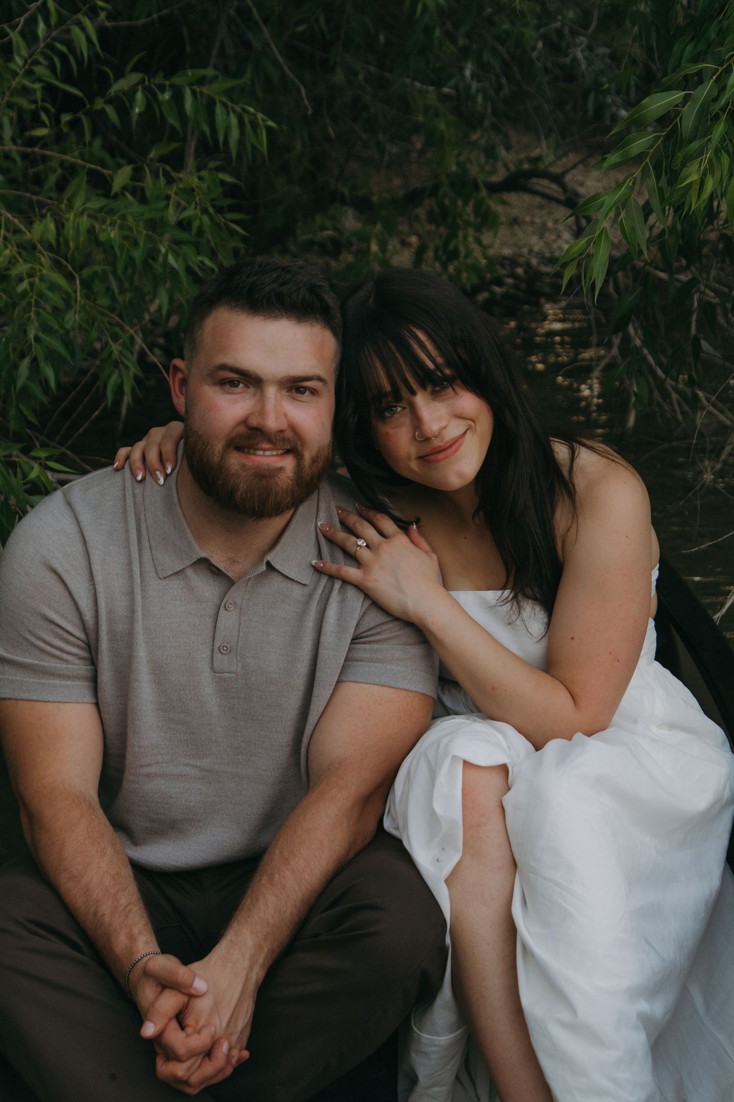 A couple sits close together in a romantic outdoor photoshoot near a lake.
