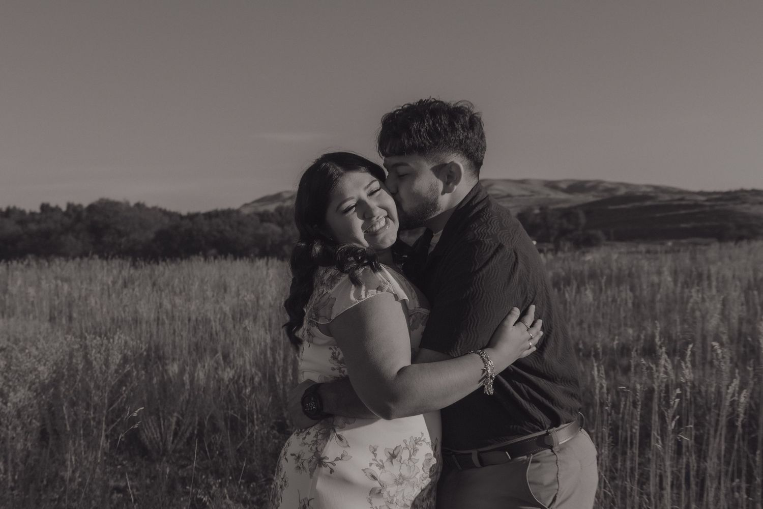 A black and white photo captures a loving embrace in a scenic field landscape.
