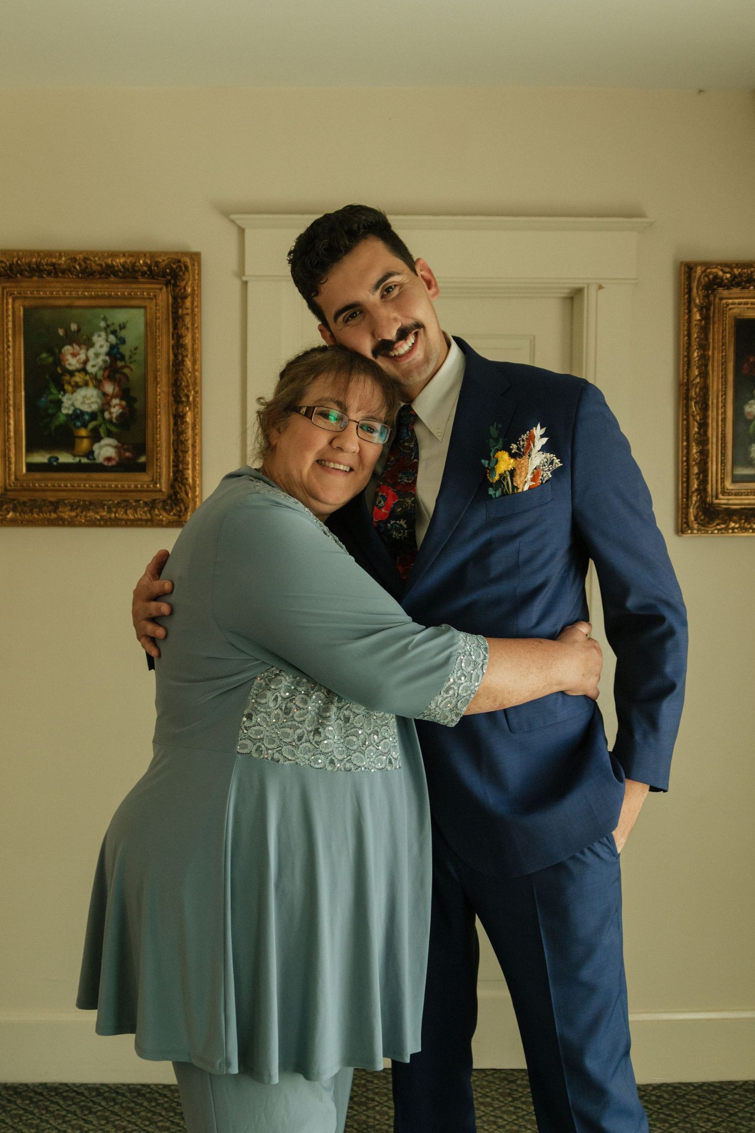 Two people share an emotional embrace, one wearing a teal dress and the other in a navy suit.