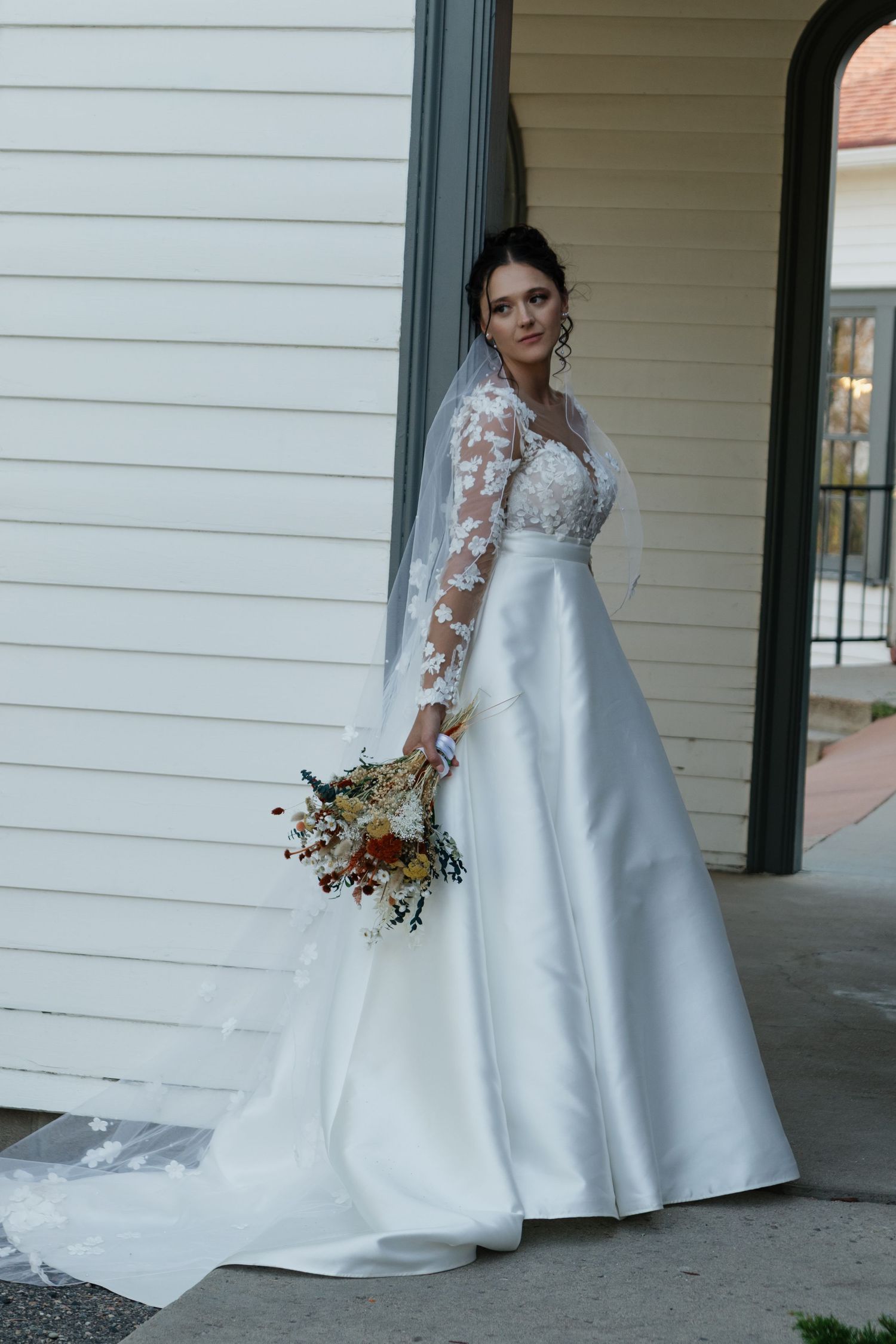 A white wedding gown with lace details and train displayed on a covered building porch.