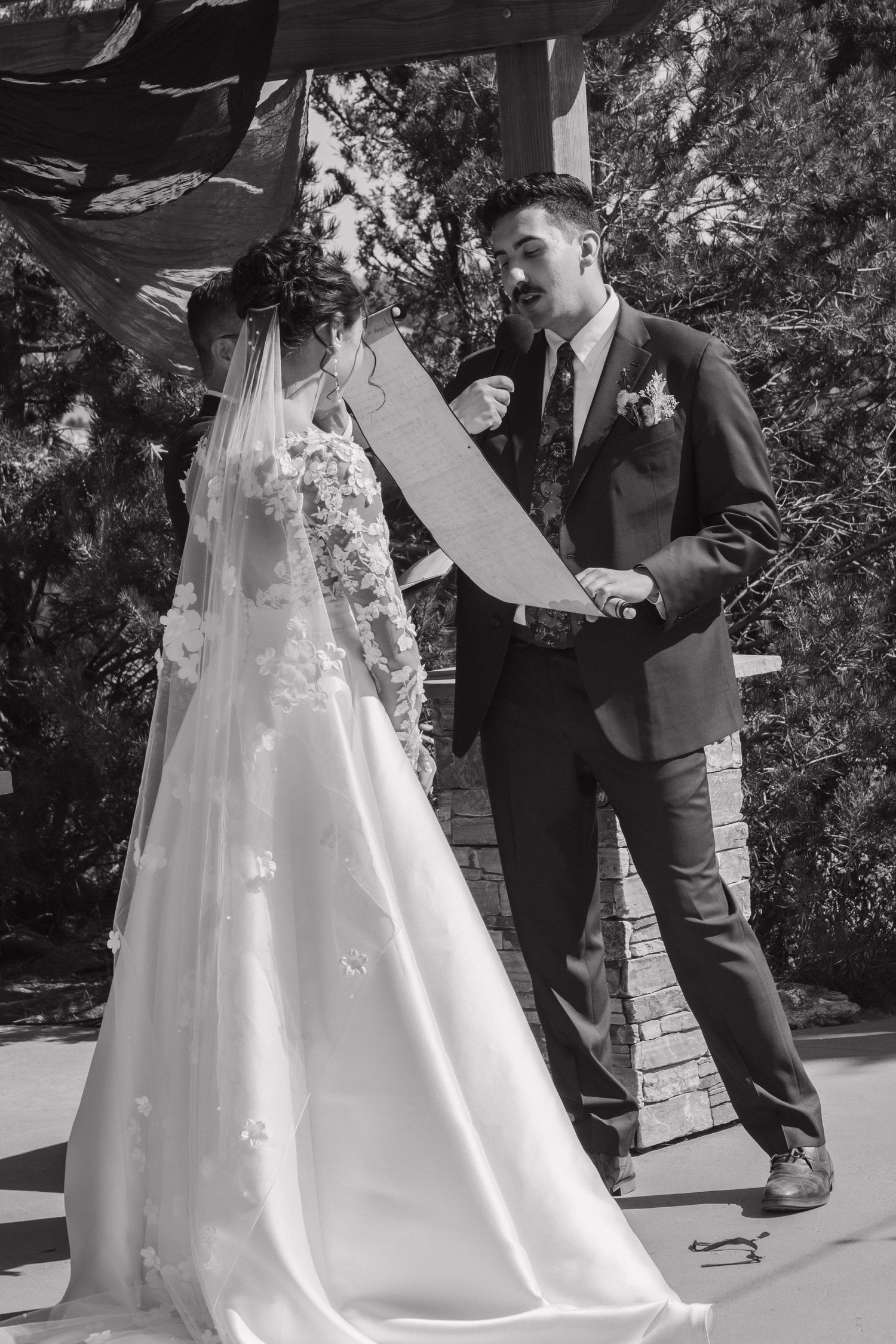 A long white wedding gown trails across dark paved stones during an outdoor ceremony.