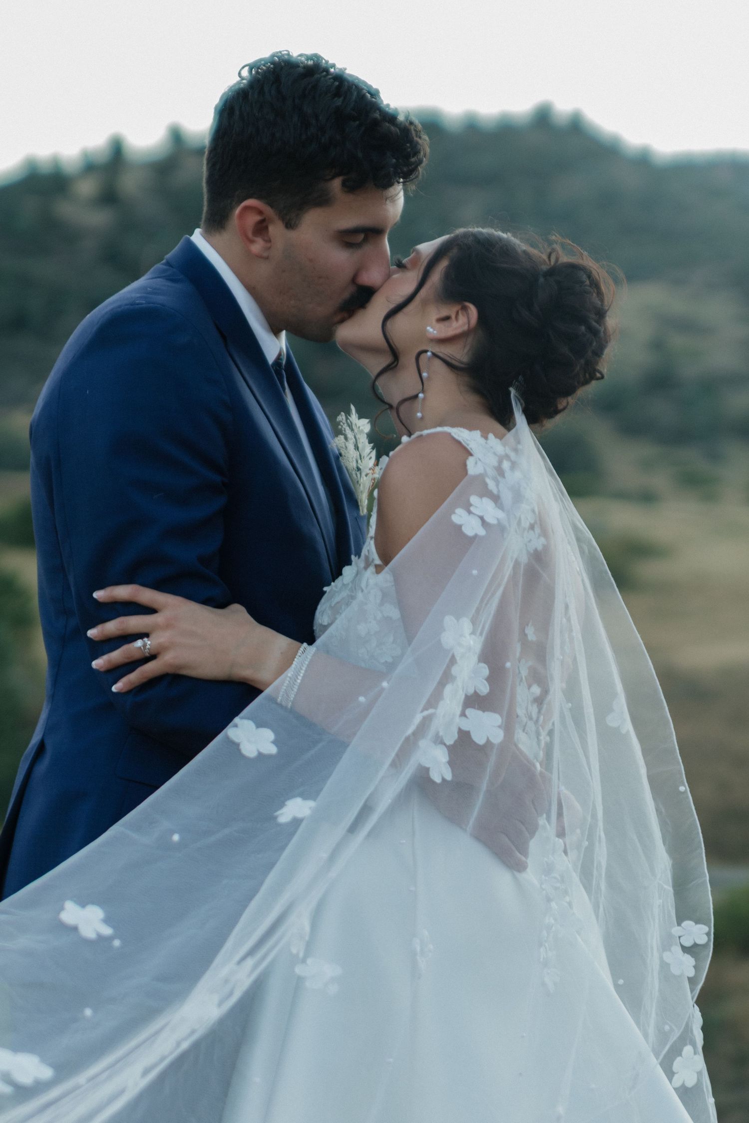 Couple embracing outdoors with flowing veil against mountain backdrop.