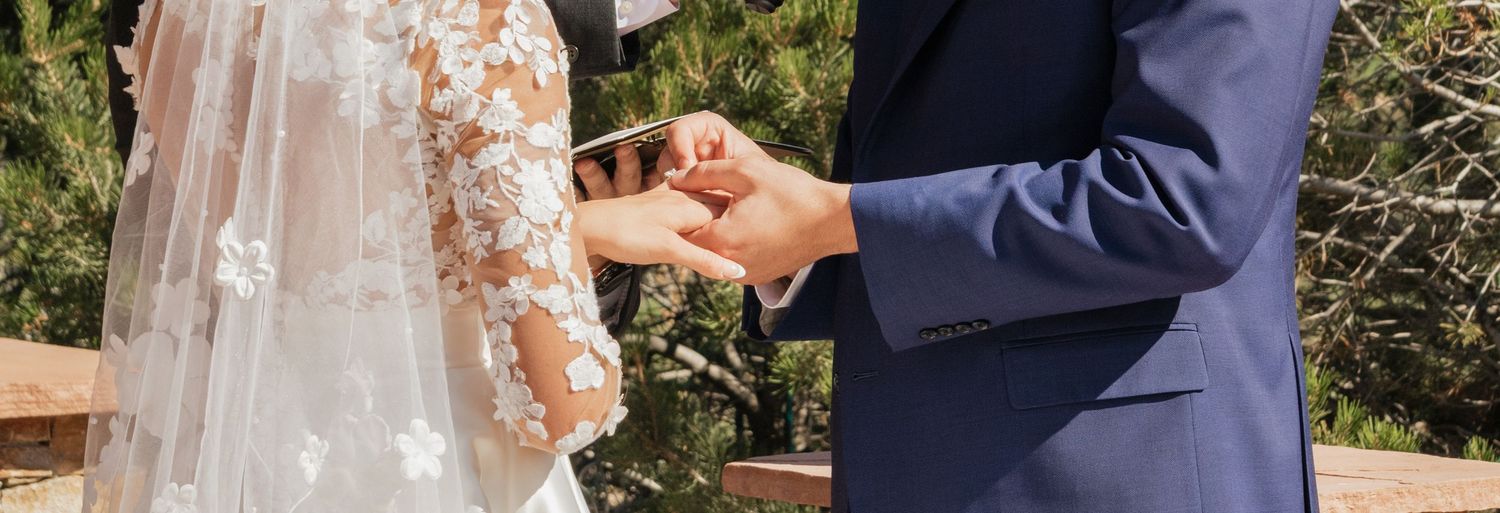 Close-up of couple exchanging rings during outdoor wedding ceremony against mountain backdrop.