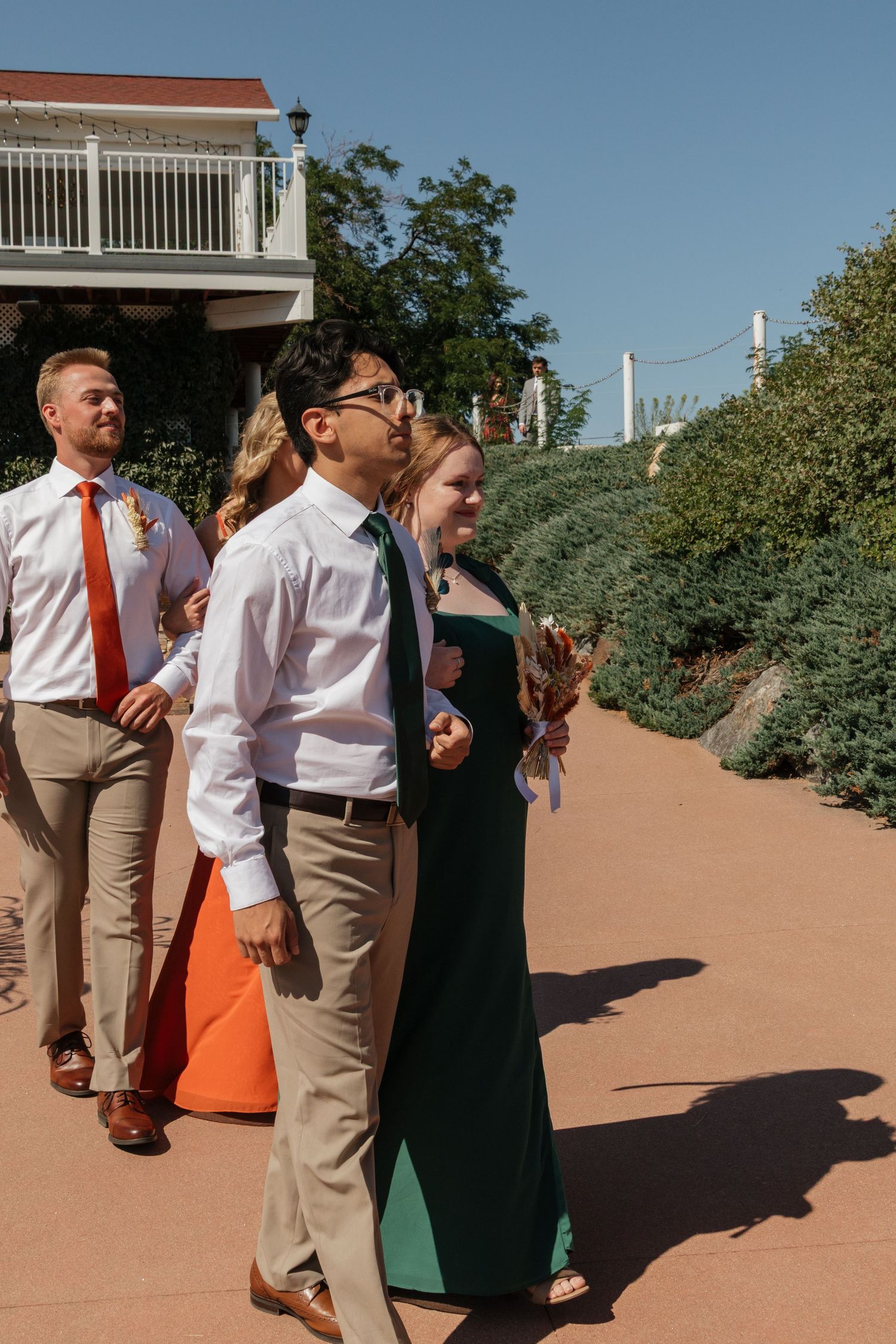 Wedding guests dressed in orange, white and khaki gather at an outdoor venue with stone pathways.