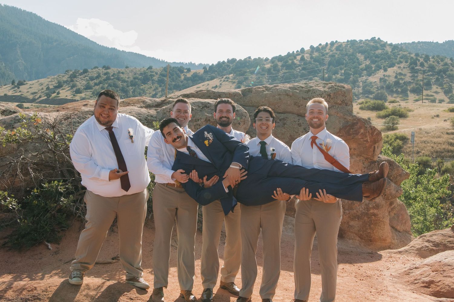 Groomsmen in khaki pants posing together on a rocky mountain overlook.