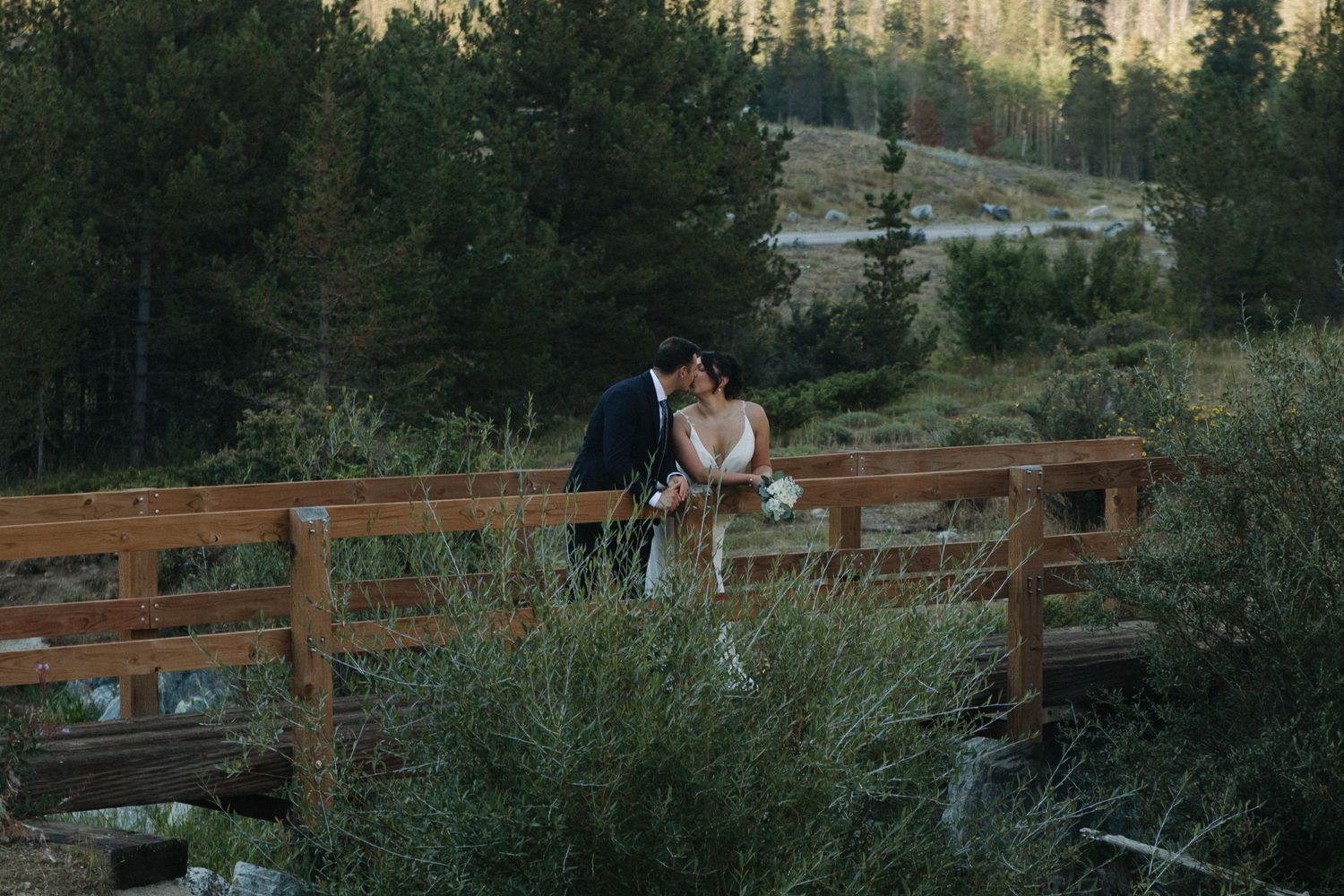 Wedding couple embraces on a rustic bridge with mountain forest landscape in background.