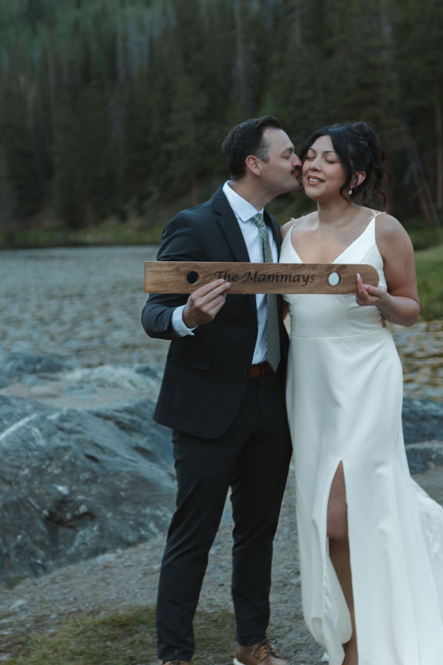 Couple shares intimate moment with wooden beam against mountain lake backdrop.