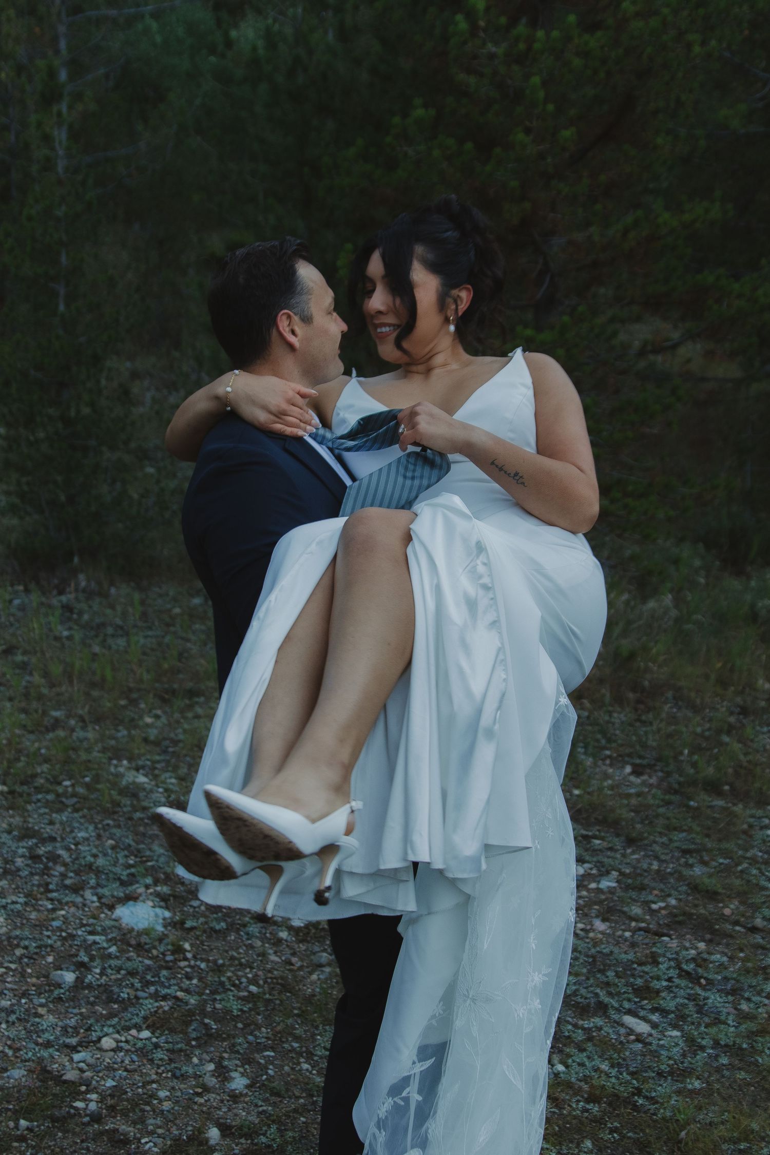 A romantic couple shares an intimate embrace in white attire against a dark forest backdrop.