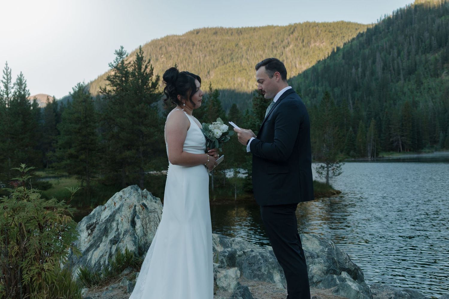 A couple shares an intimate moment during their wedding ceremony against a scenic mountain lake backdrop.