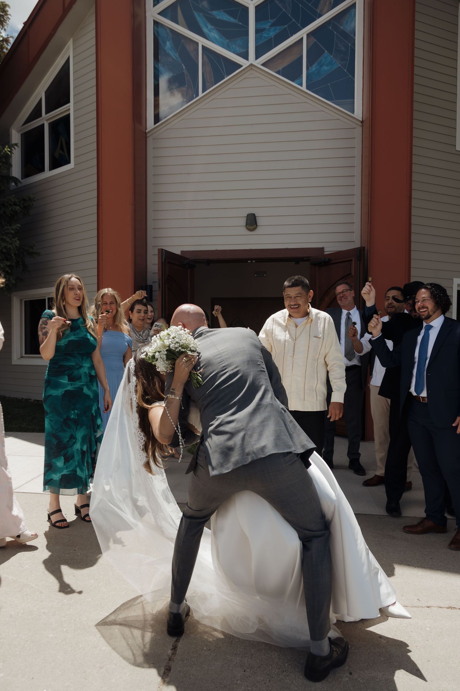 Groom dipping bride for a kiss outside church entrance surrounded by cheering wedding guests.