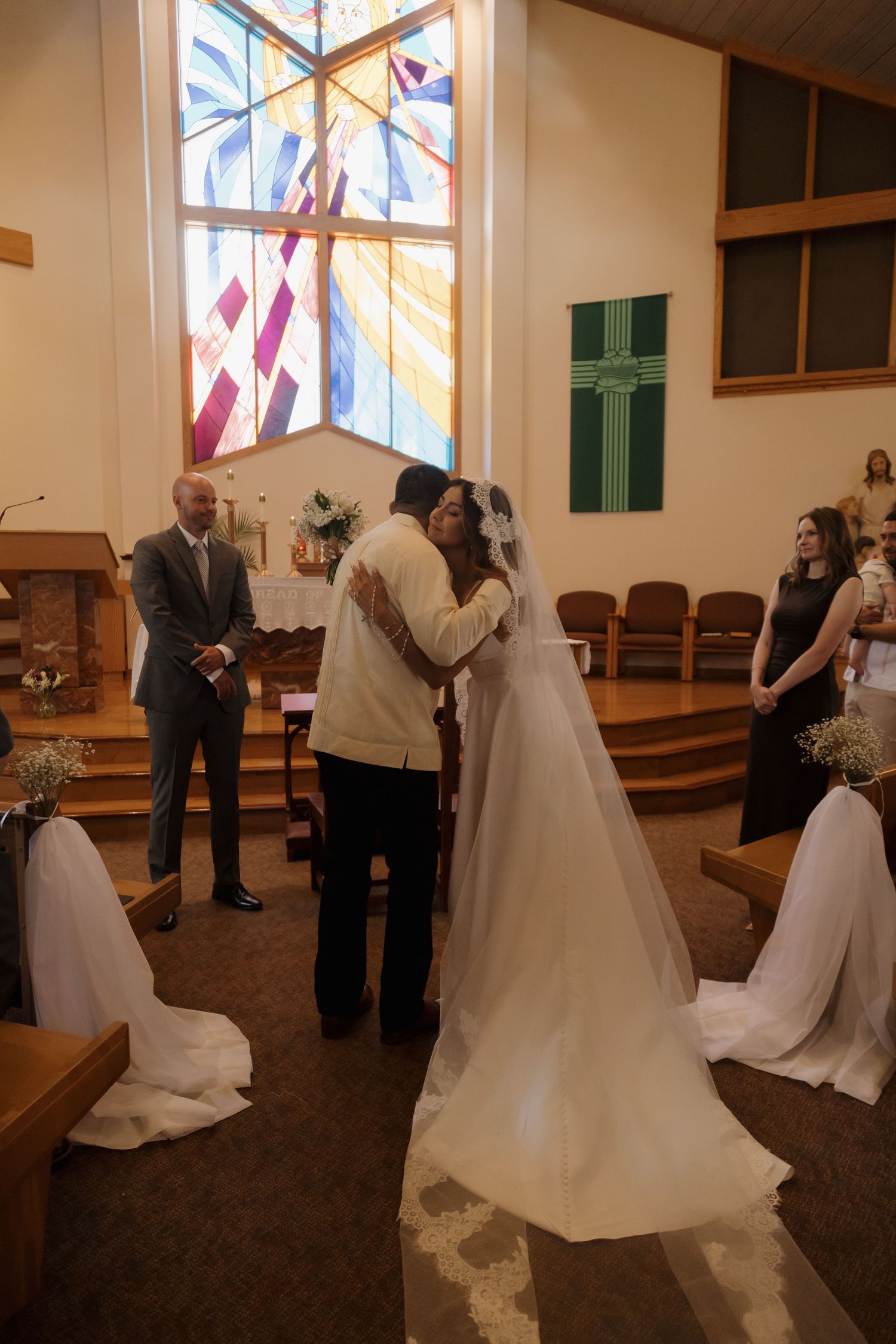 Bride and groom share their first kiss at the altar beneath a colorful stained glass window in a Catholic church.