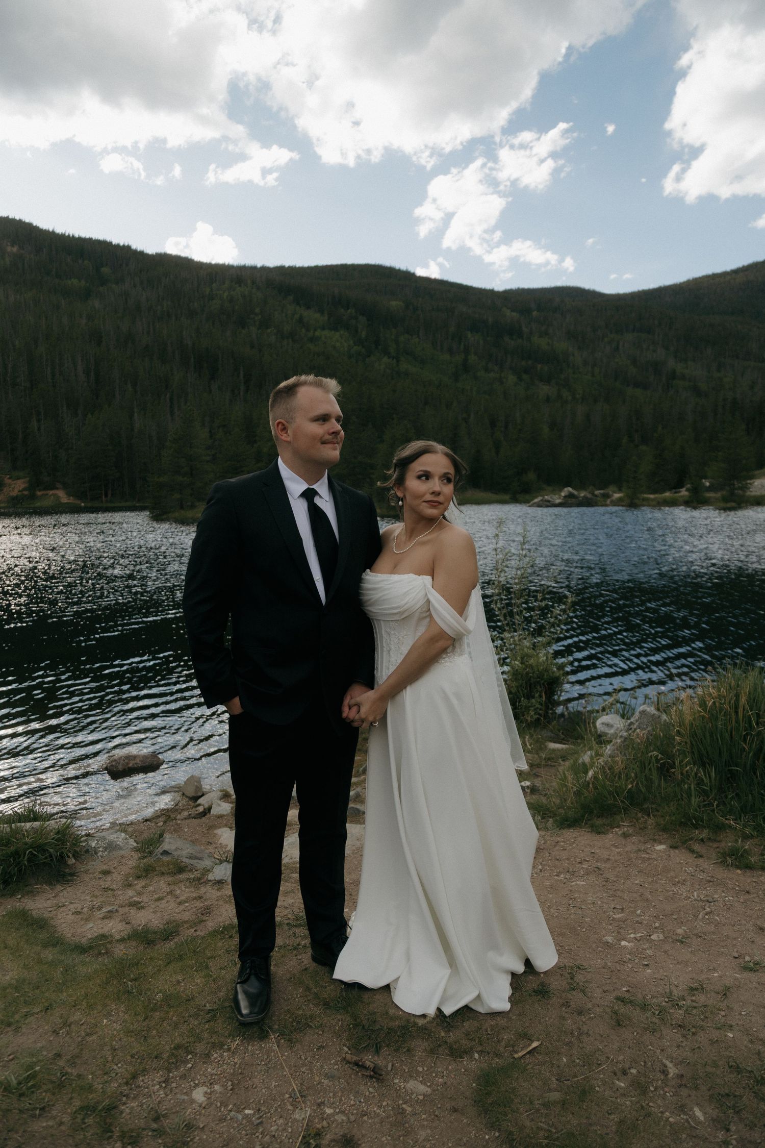 Bride and groom hold hands by a mountain lake, she in a white off-shoulder gown, he in a black suit.