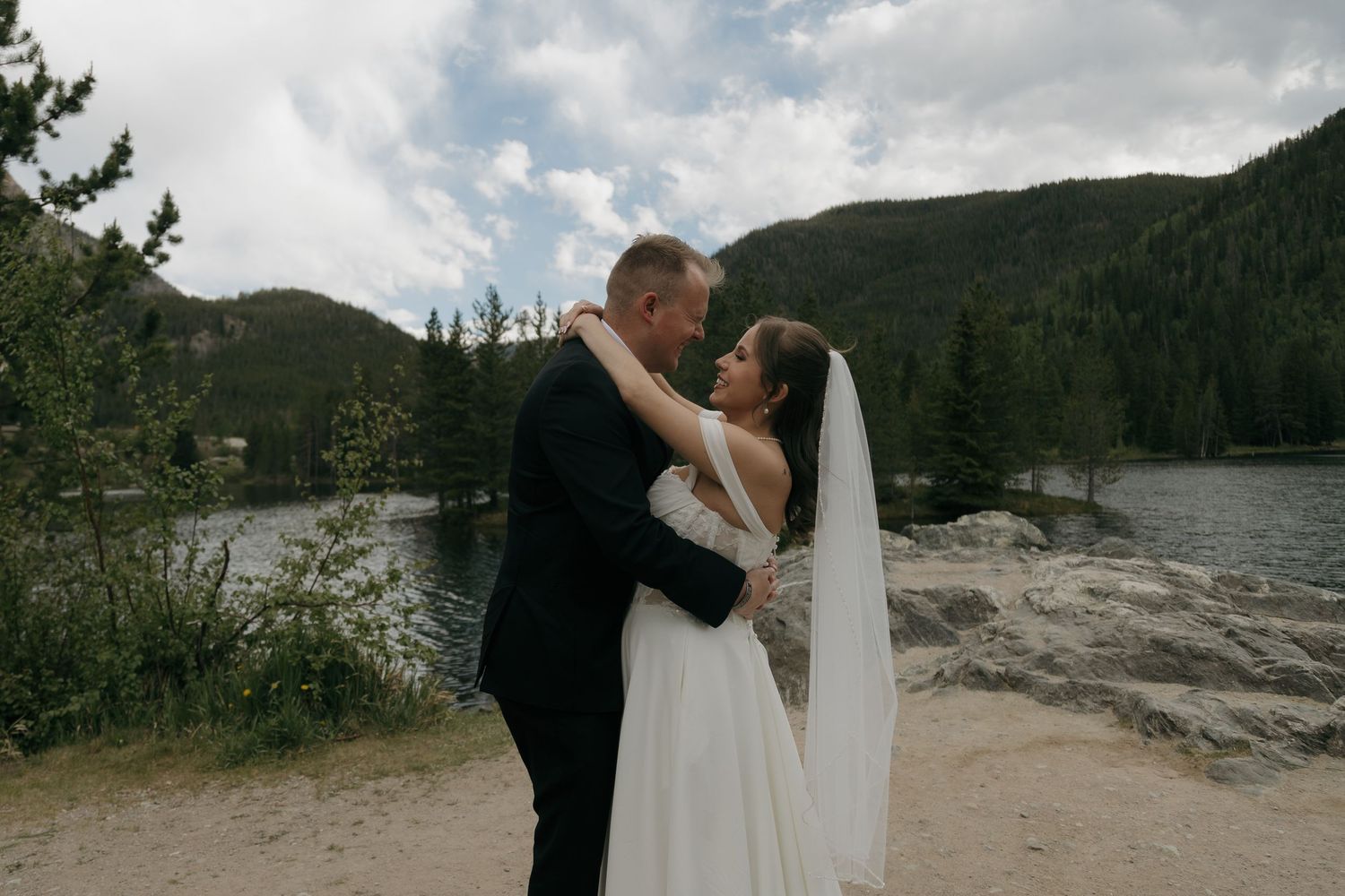 Bride and groom share a romantic embrace near a mountain lake surrounded by pine trees on their wedding day.