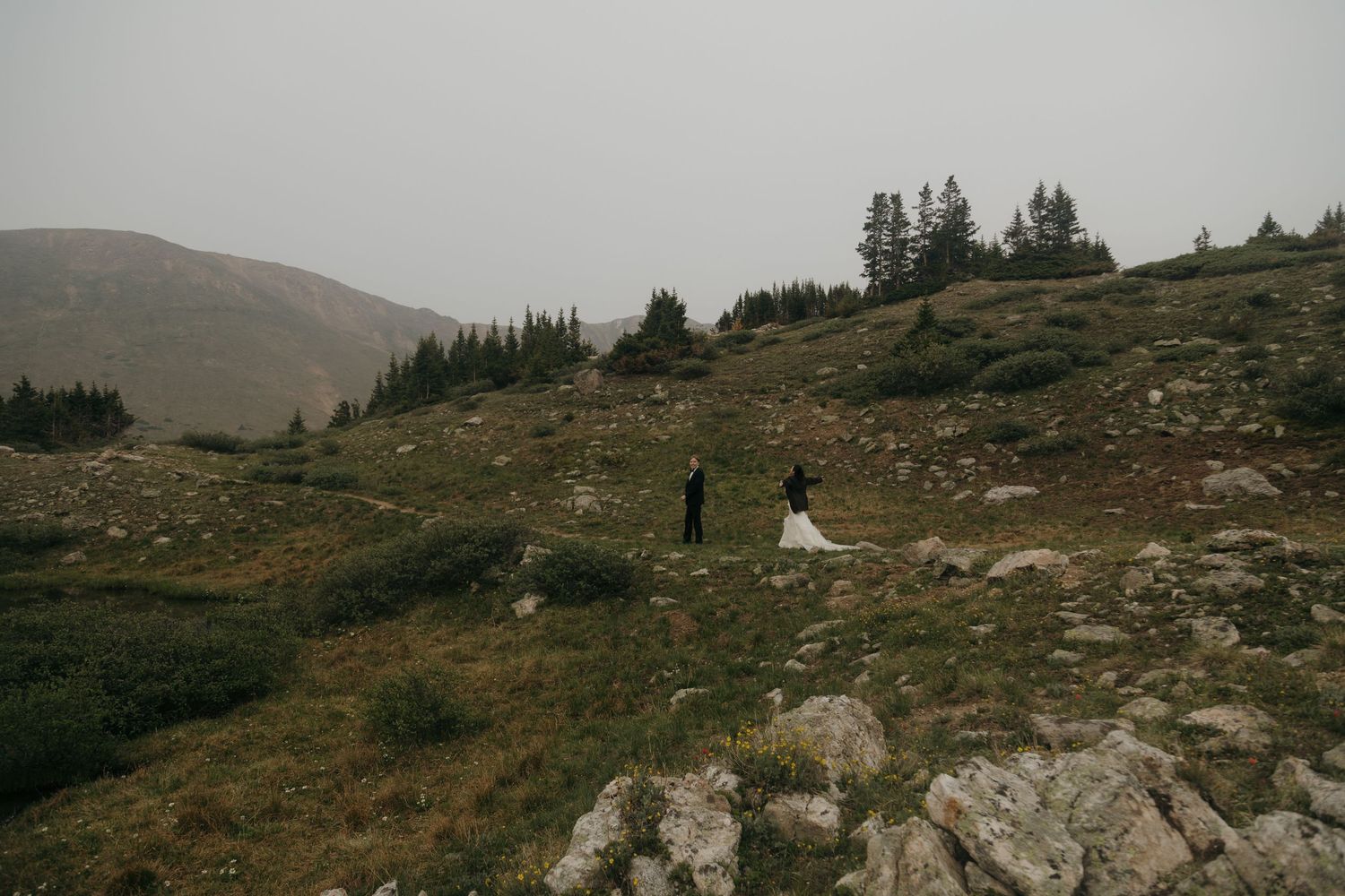 Bride and groom stand together on a misty mountain hillside surrounded by evergreen trees during their elopement.