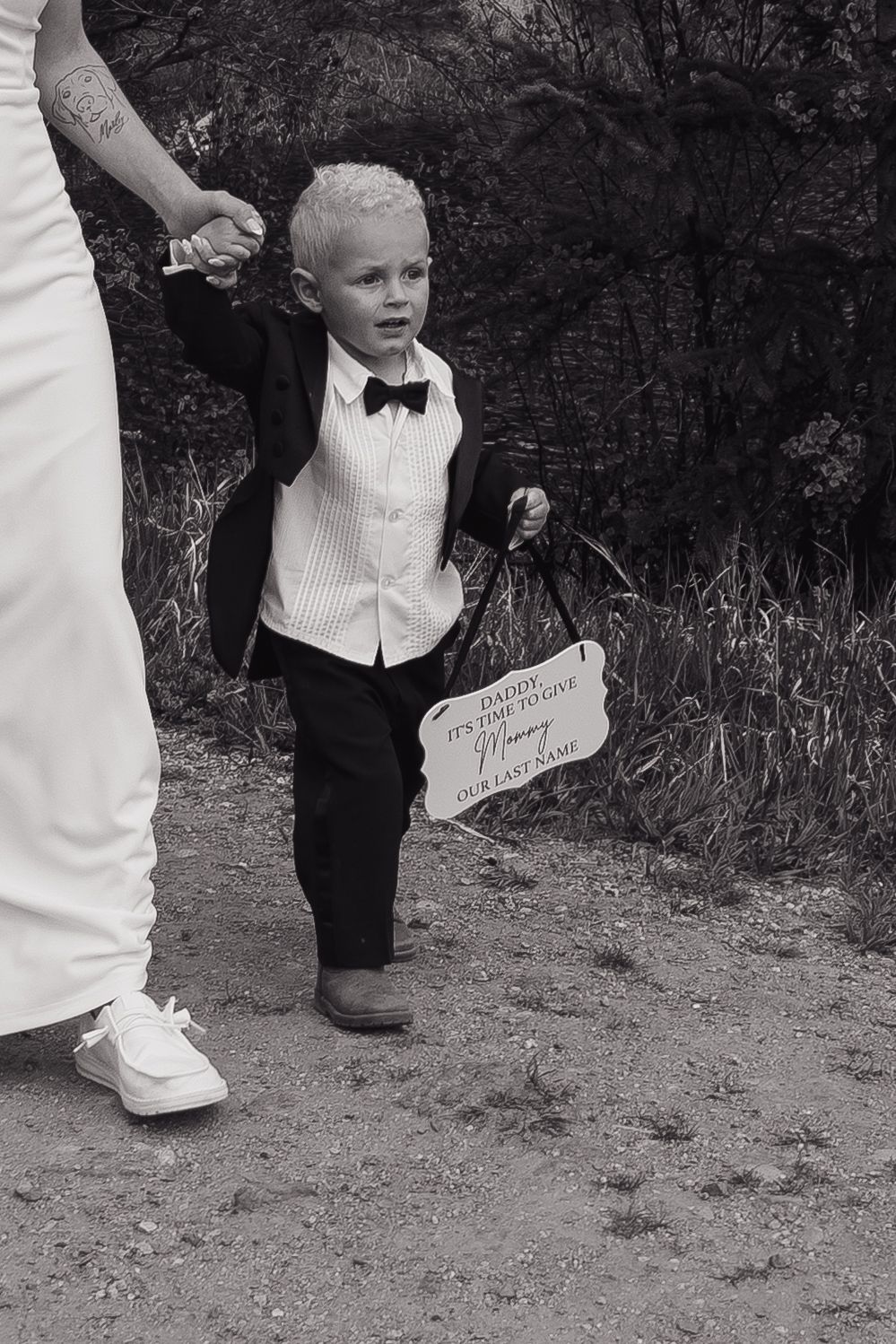 A young ring bearer in a tuxedo holds a wedding sign while walking with the bride on a dirt path.