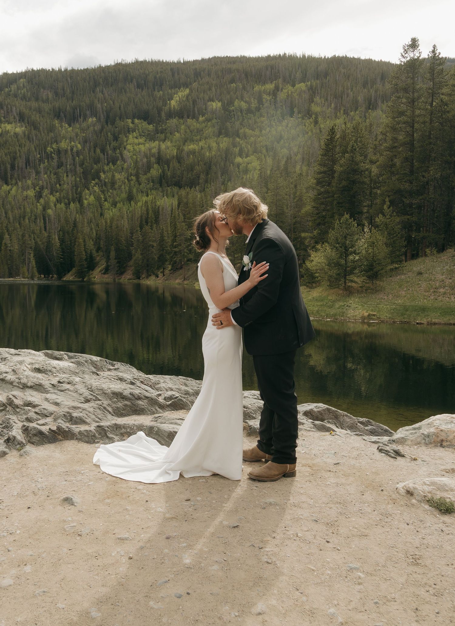 A bride and groom share a first kiss on rocky terrain beside a serene mountain lake.