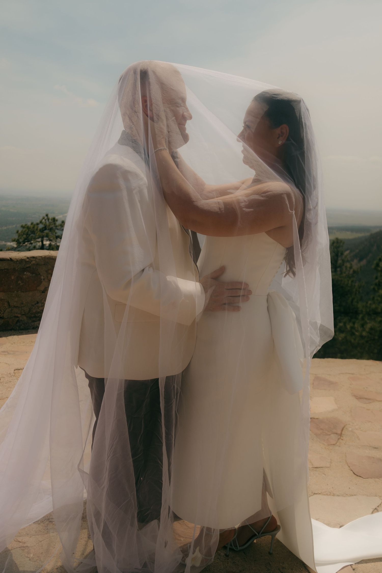 Bride and groom share intimate moment beneath flowing wedding veil on mountain overlook.