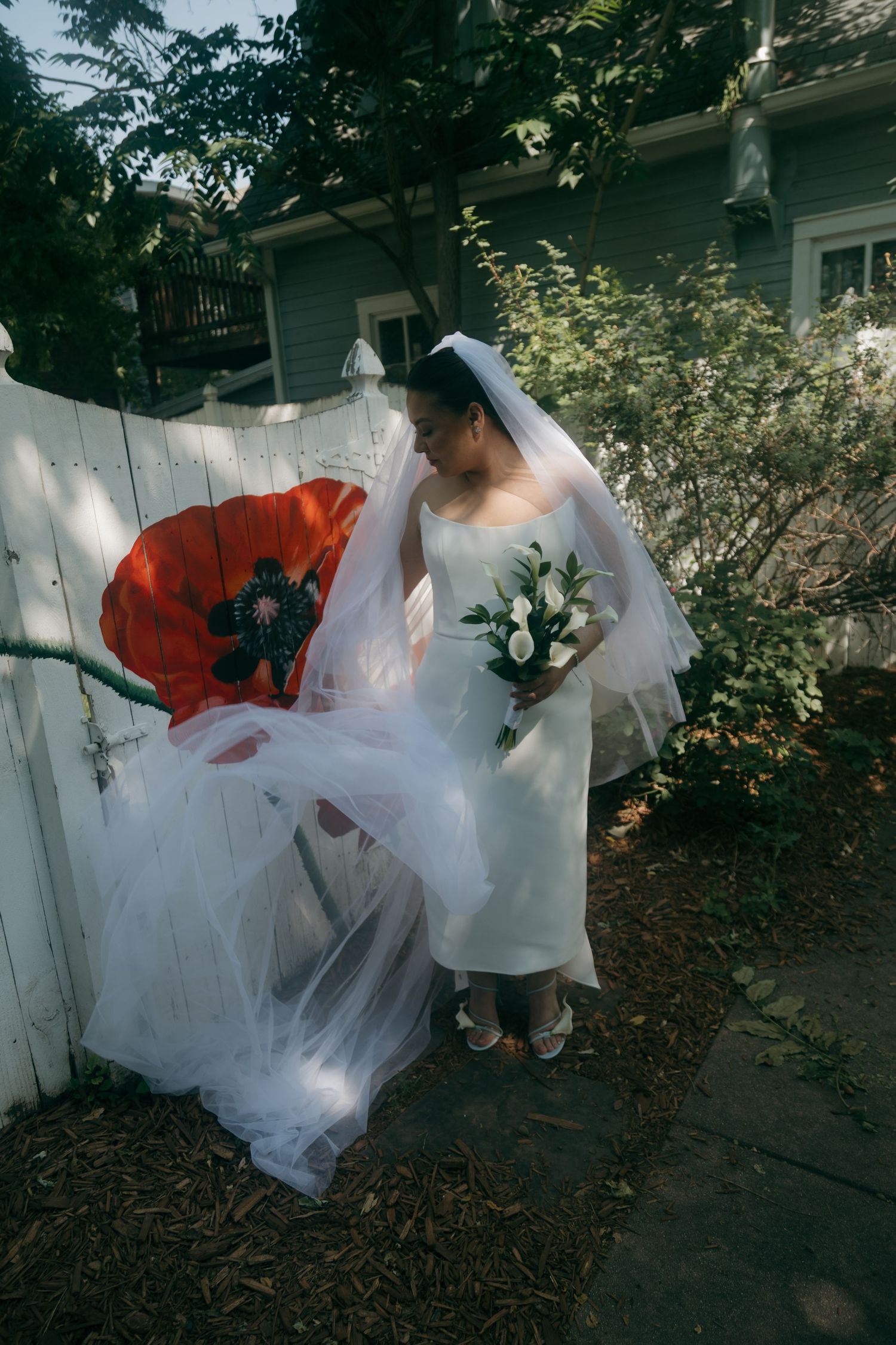 Bride in white gown and veil holds bouquet beside garden fence.