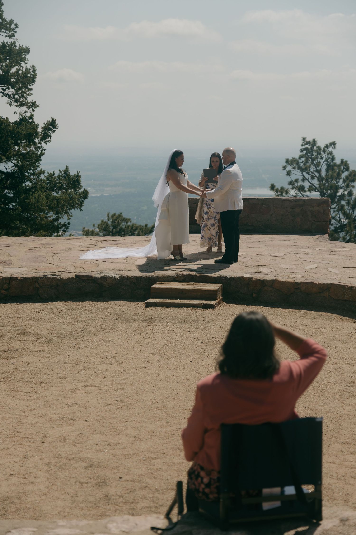 Outdoor mountain wedding ceremony with bride and groom exchanging vows at scenic overlook while guest watches from chair.